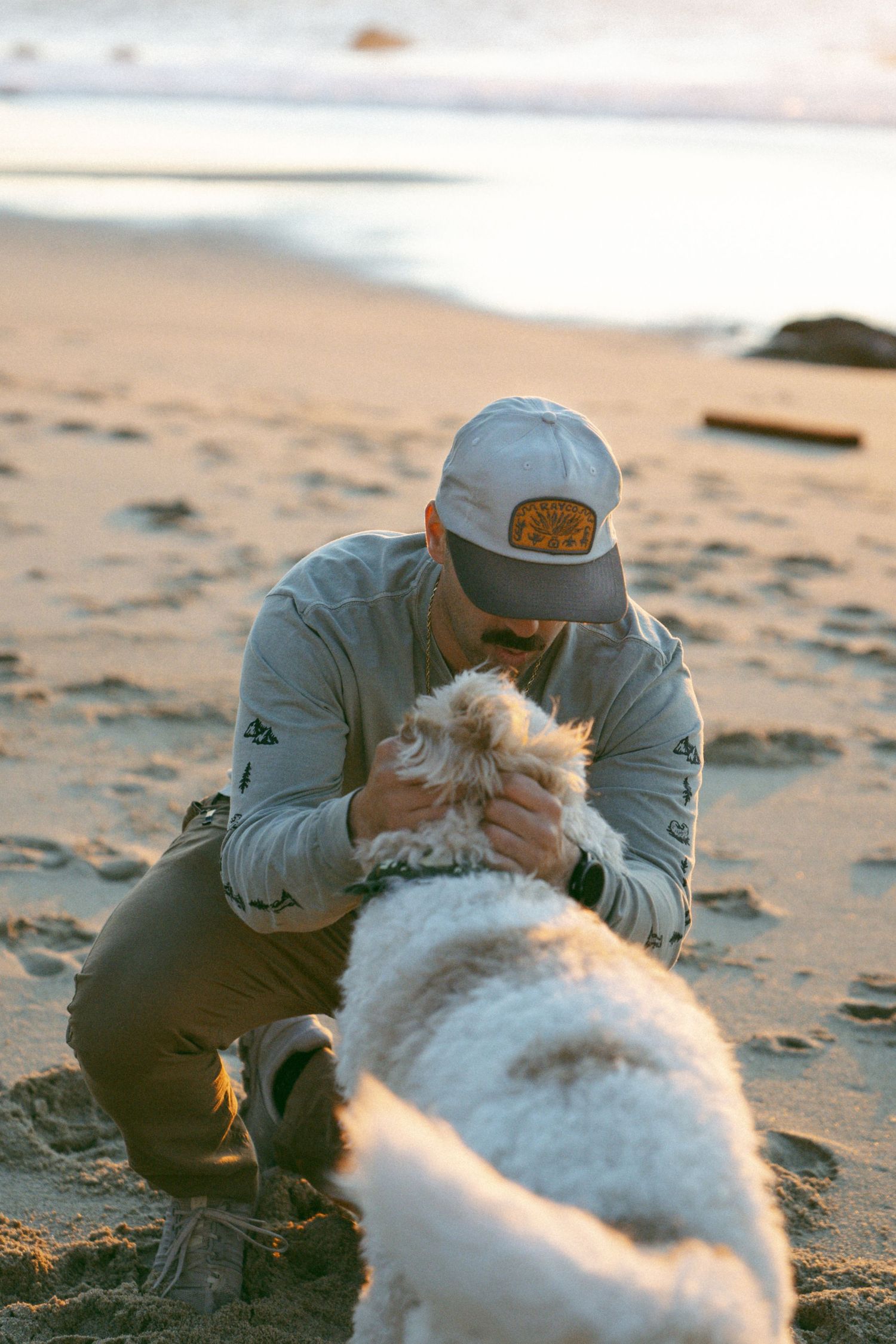 Person in casual wear plays with a fluffy white dog on a sandy beach at sunset.