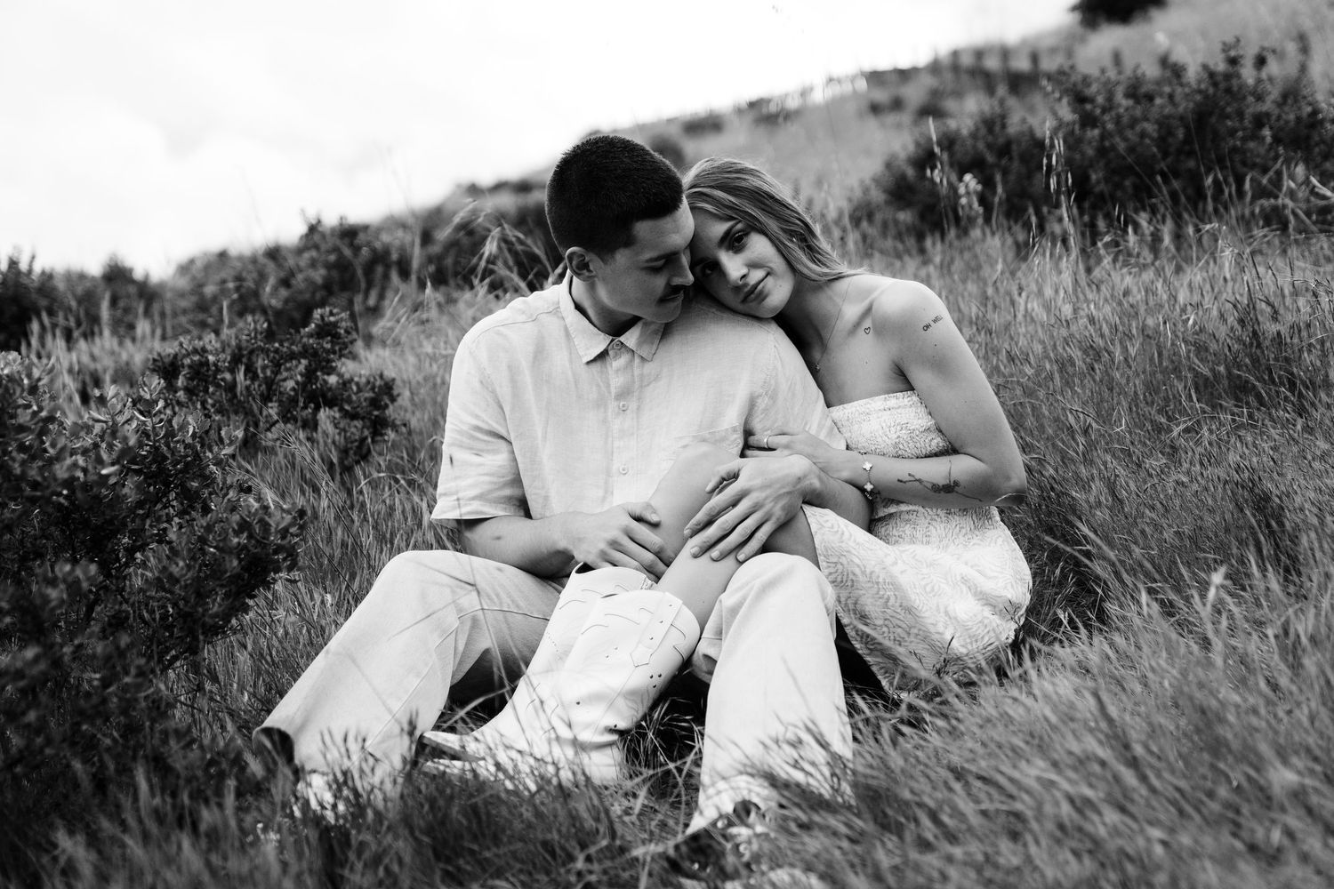 A couple shares an intimate black and white moment together while sitting in a grassy field on a hillside in Big Sur California.