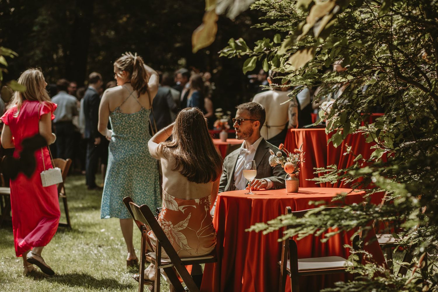 Guests enjoy an outdoor garden party with red tablecloths and colorful summer dresses on a sunny afternoon.