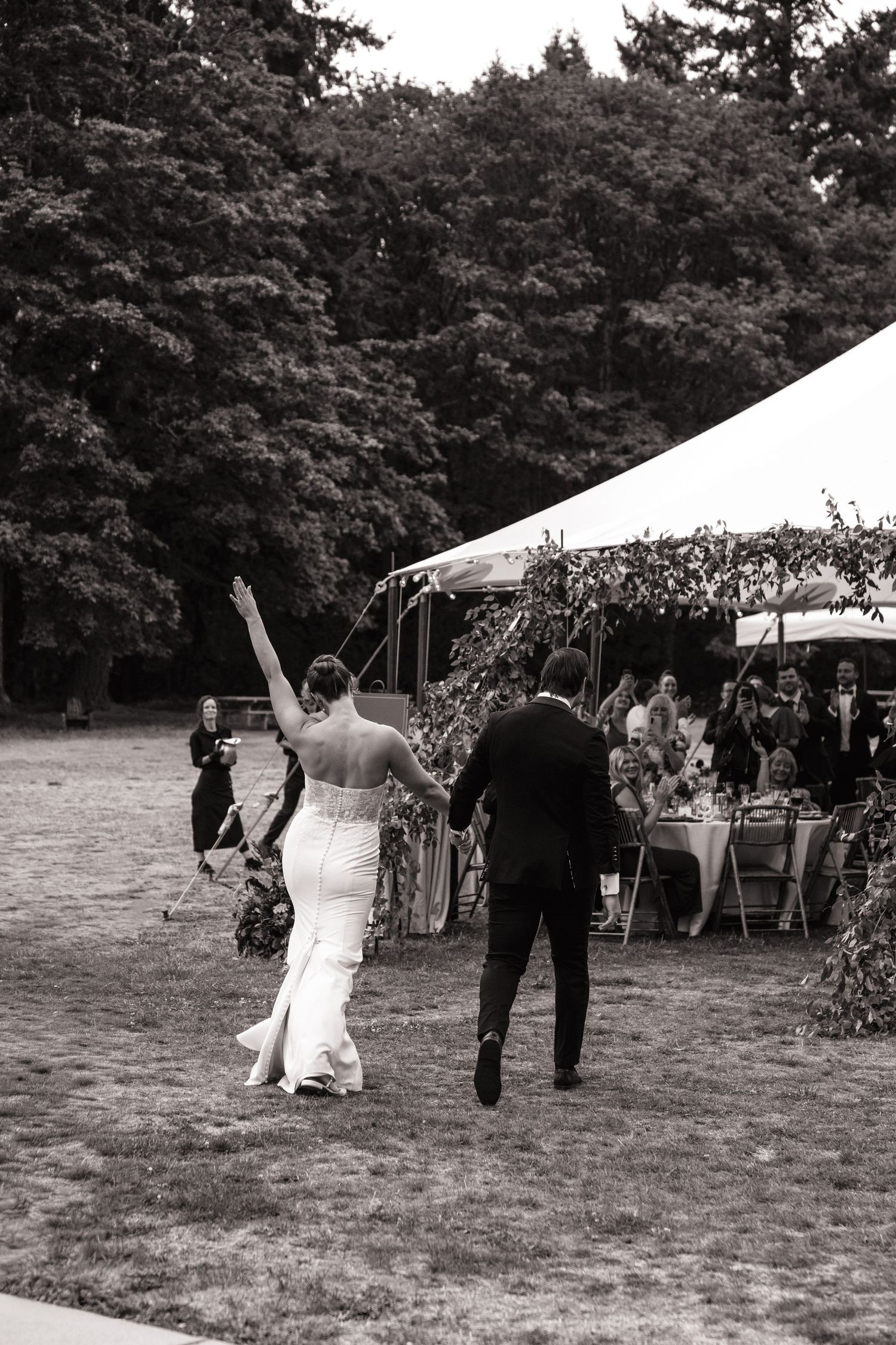 Black and white photo of wedding guests walking across a grassy field with tents in the background.