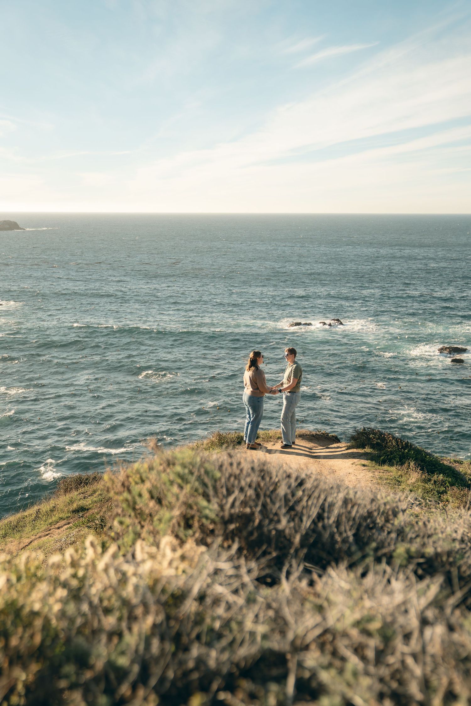 Two silhouettes stand together on a scenic coastal cliff overlooking the vast ocean at sunset.