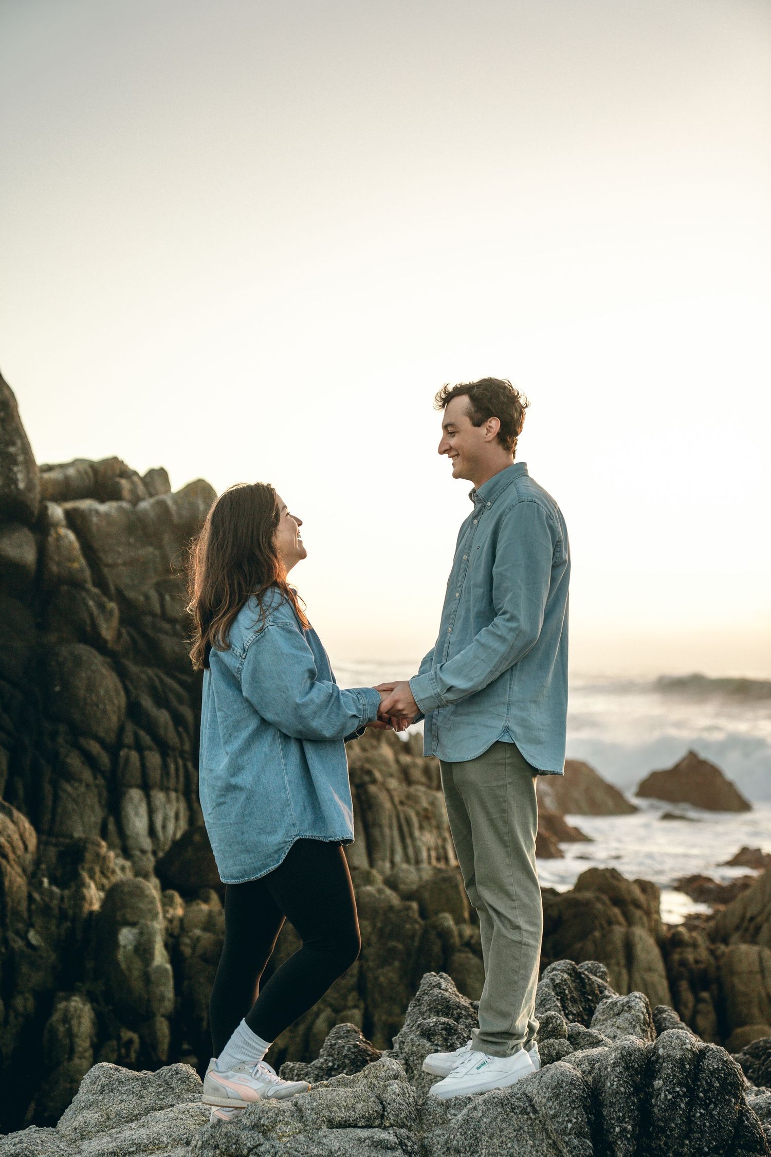 Couple in blue denim sharing joyful moments on rocky coastal cliffs at dusk.