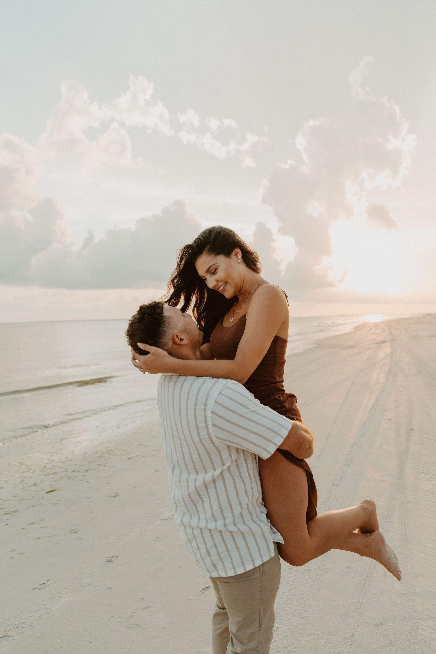 A romantic couple shares an intimate moment on a beach at sunset with soft clouds in the sky.