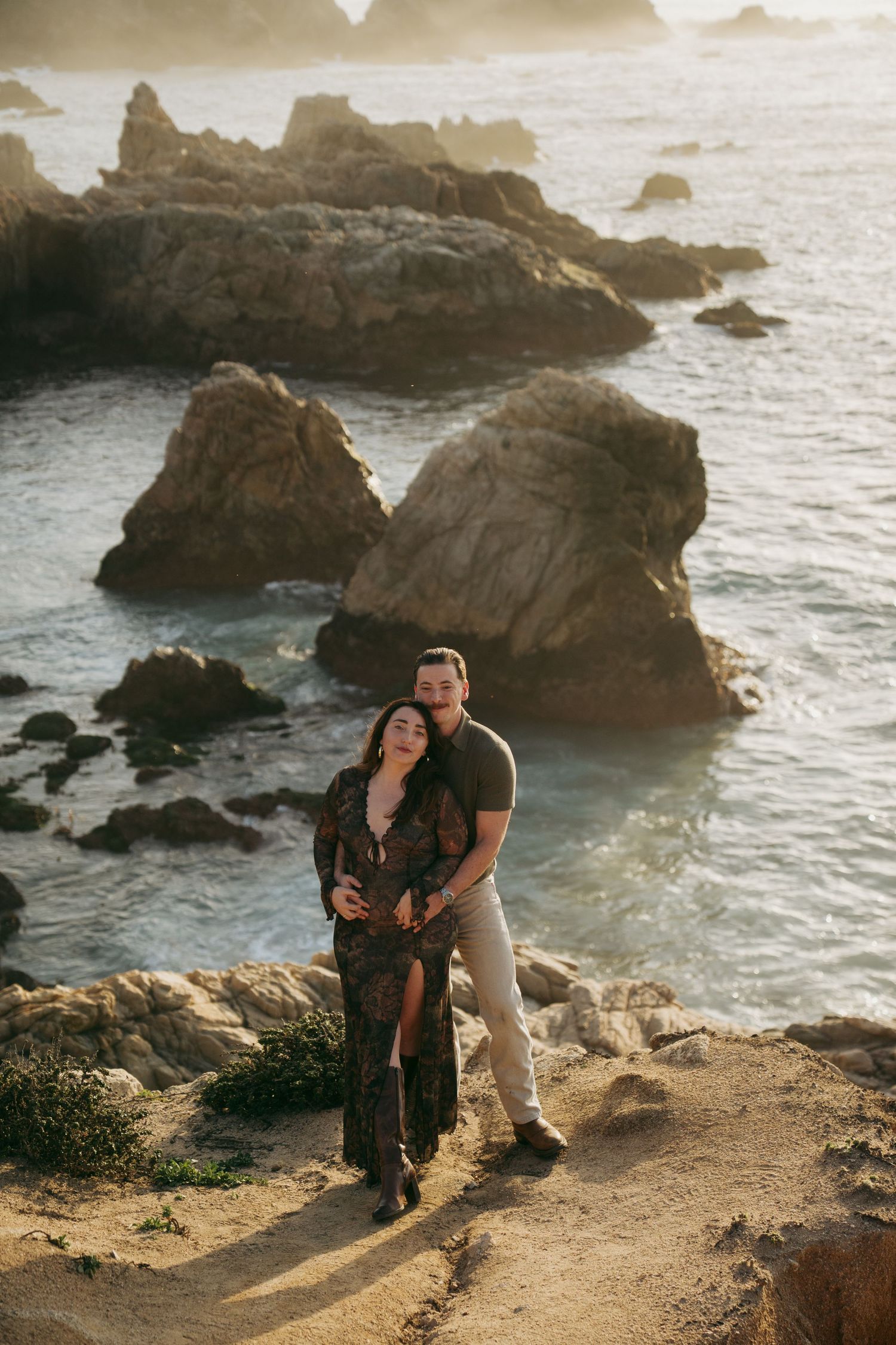 Couple embraces on a rocky coastal cliff overlooking the ocean at sunset with dramatic rock formations in background.
