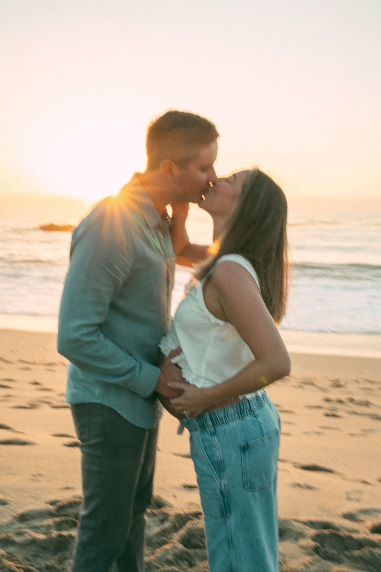 Couple embracing and kissing on sandy beach during golden hour with ocean waves in background.