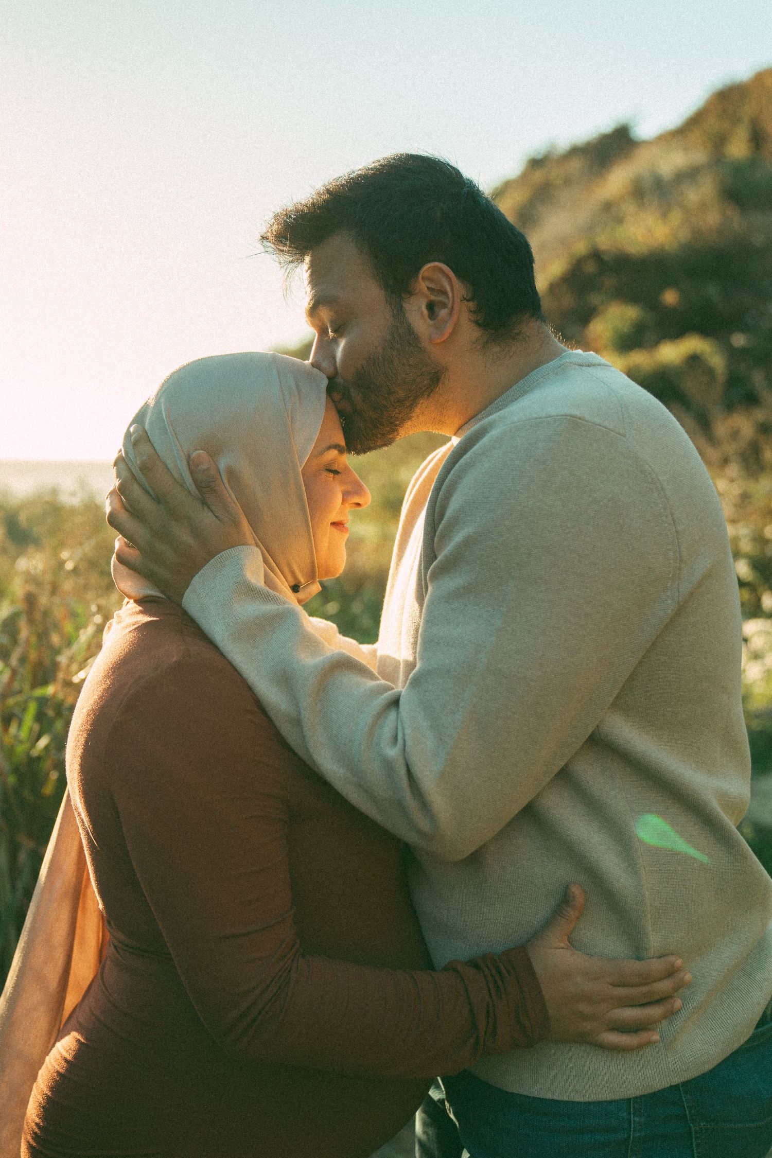 A couple shares an intimate moment outdoors during golden hour, with the woman wearing a hijab and light glowing behind them.