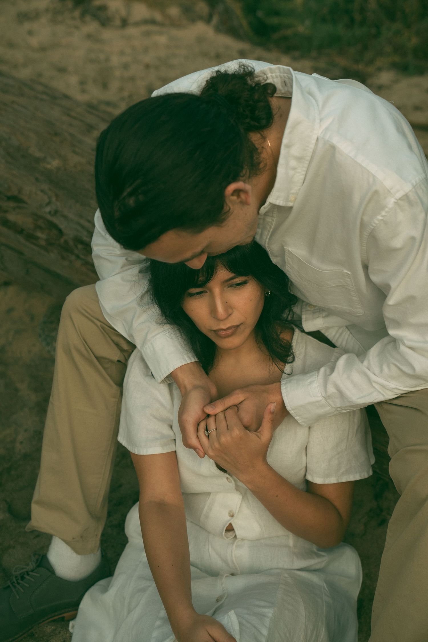 A romantic couple embraces tenderly outdoors, both wearing white clothing in a vintage-style intimate portrait.