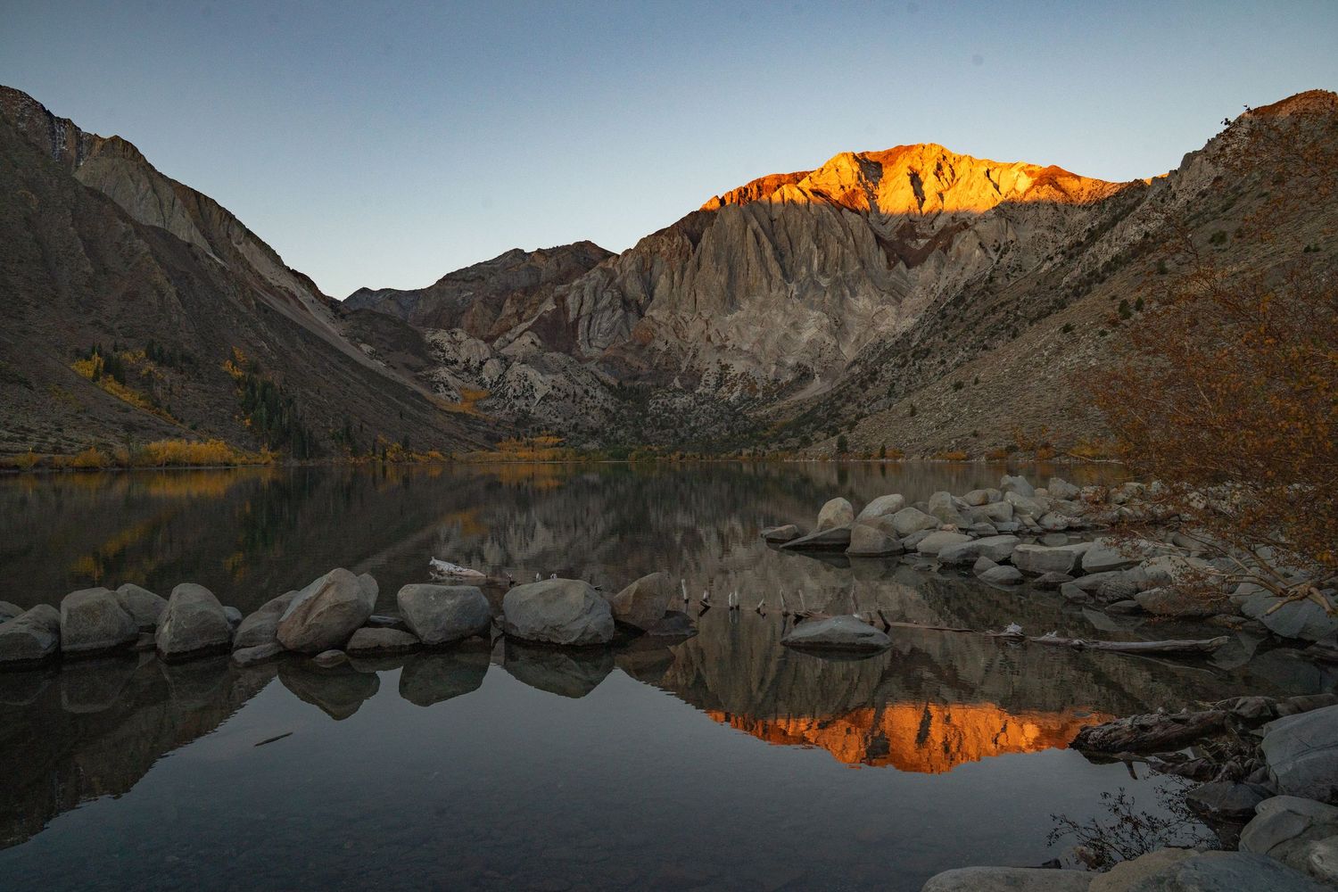 Panoramic mountain landscape with golden sunlit peaks reflected in a calm alpine lake at sunset.