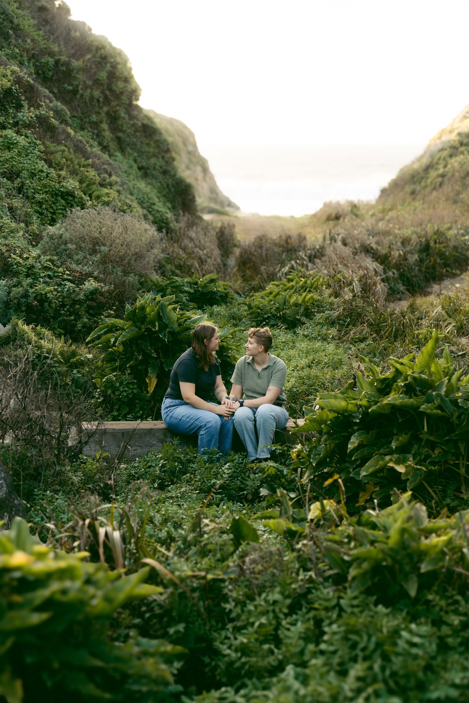 Two people sit together on a hillside surrounded by lush greenery at sunset.