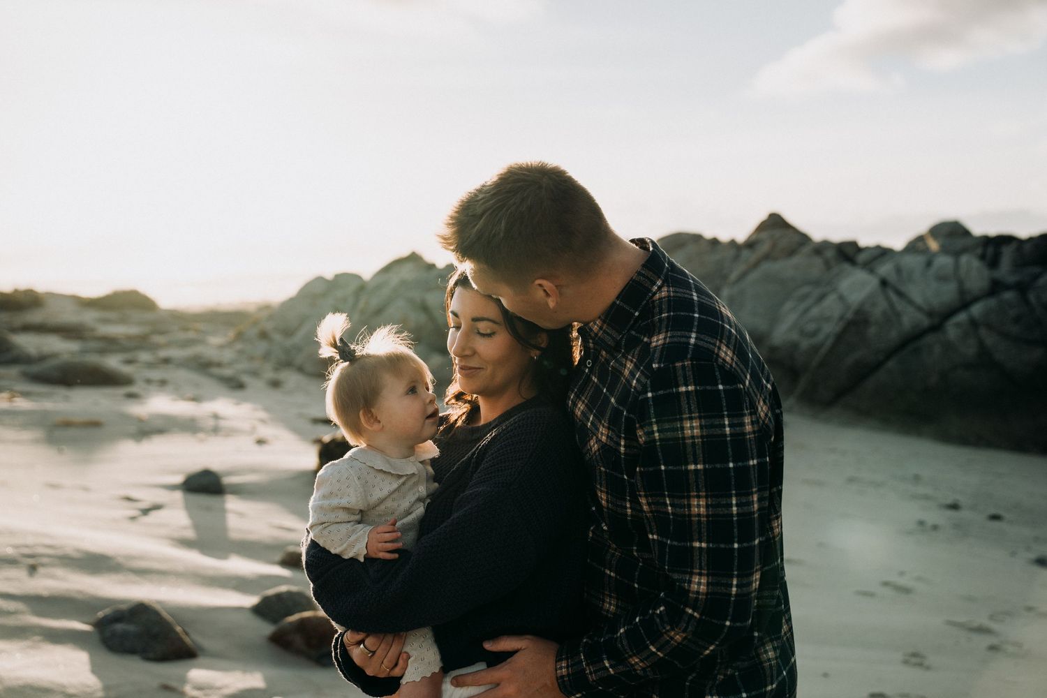 A couple shares a tender moment on the beach at sunset with their baby in a plaid shirt and casual attire.