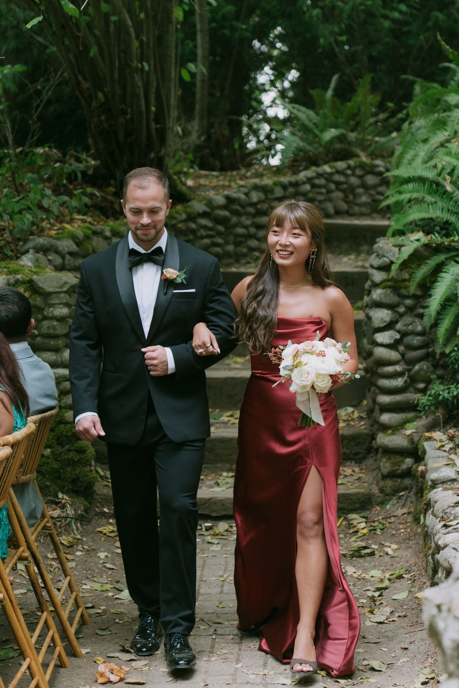 Wedding party members walk along a stone wall path surrounded by lush greenery.