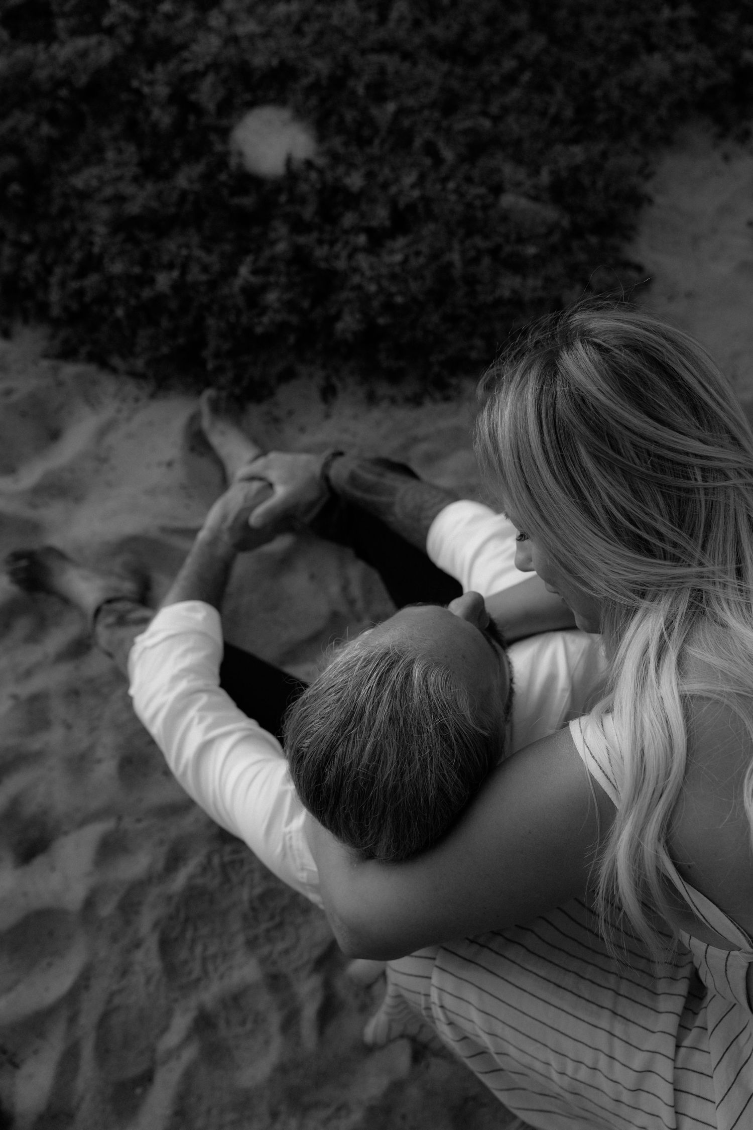 A tender black and white moment captured from above as two people embrace while sitting together on sand.