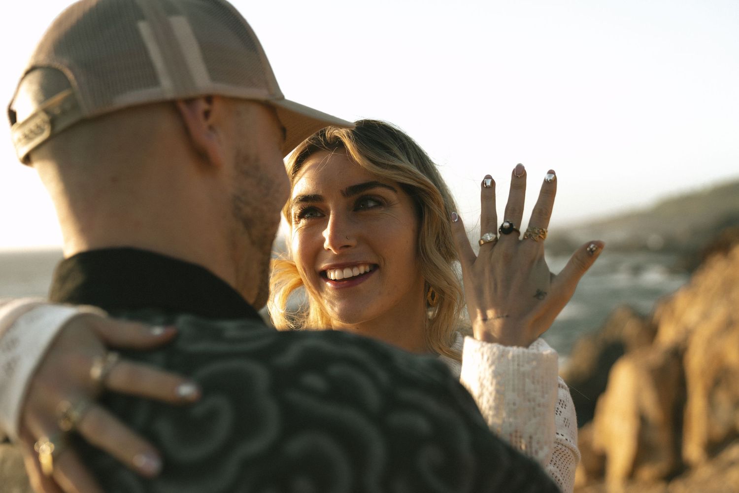 Smiling woman shows off engagement ring while embracing partner outdoors at golden hour.
