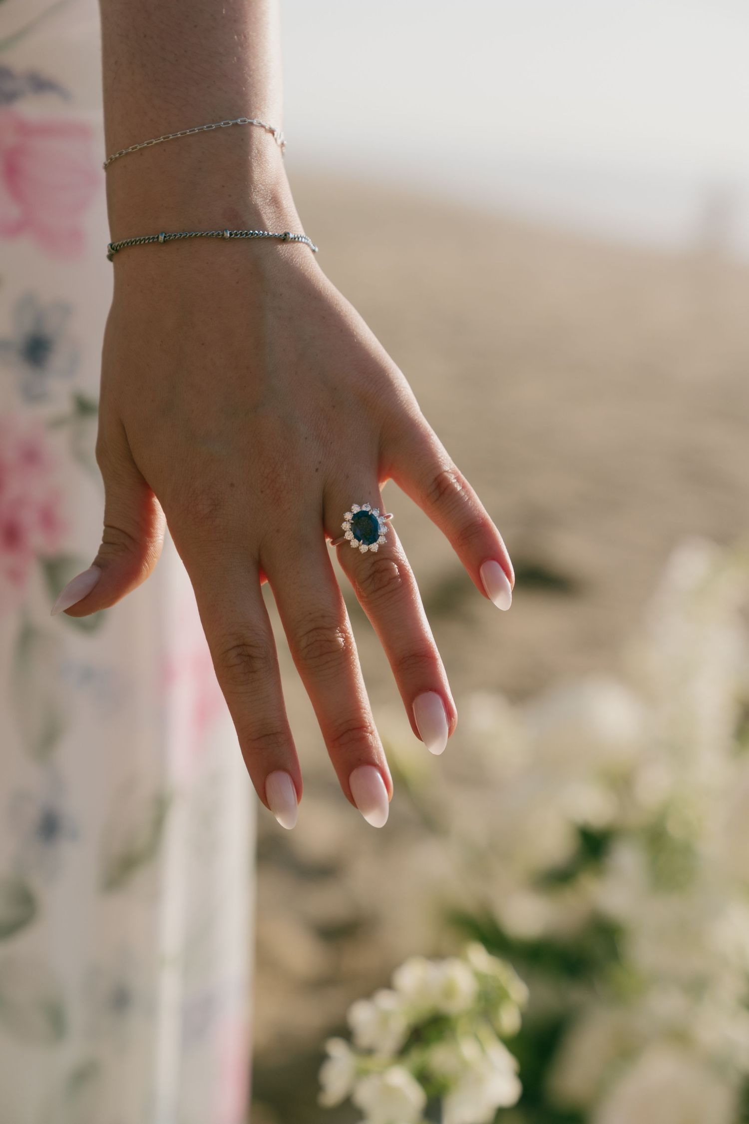 Close-up of a hand wearing a sapphire halo ring and delicate bracelet, with a blurred floral background.