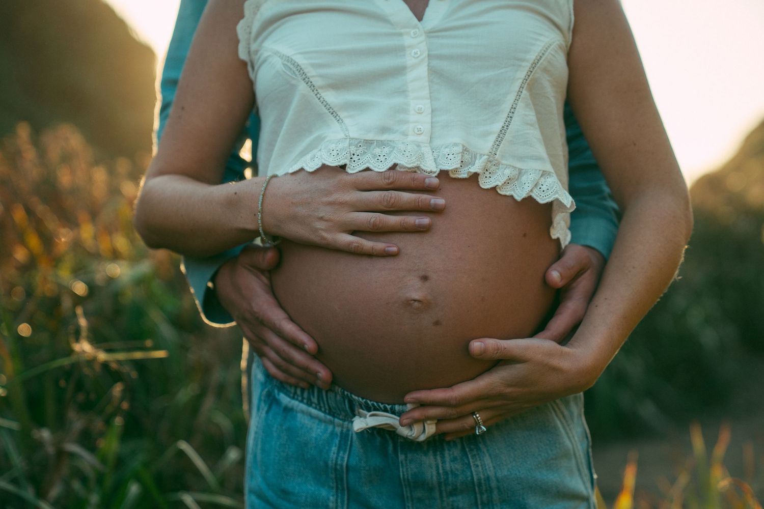 A pregnant belly is cradled by hands wearing turquoise bracelets against a sunset-lit field of wildflowers.