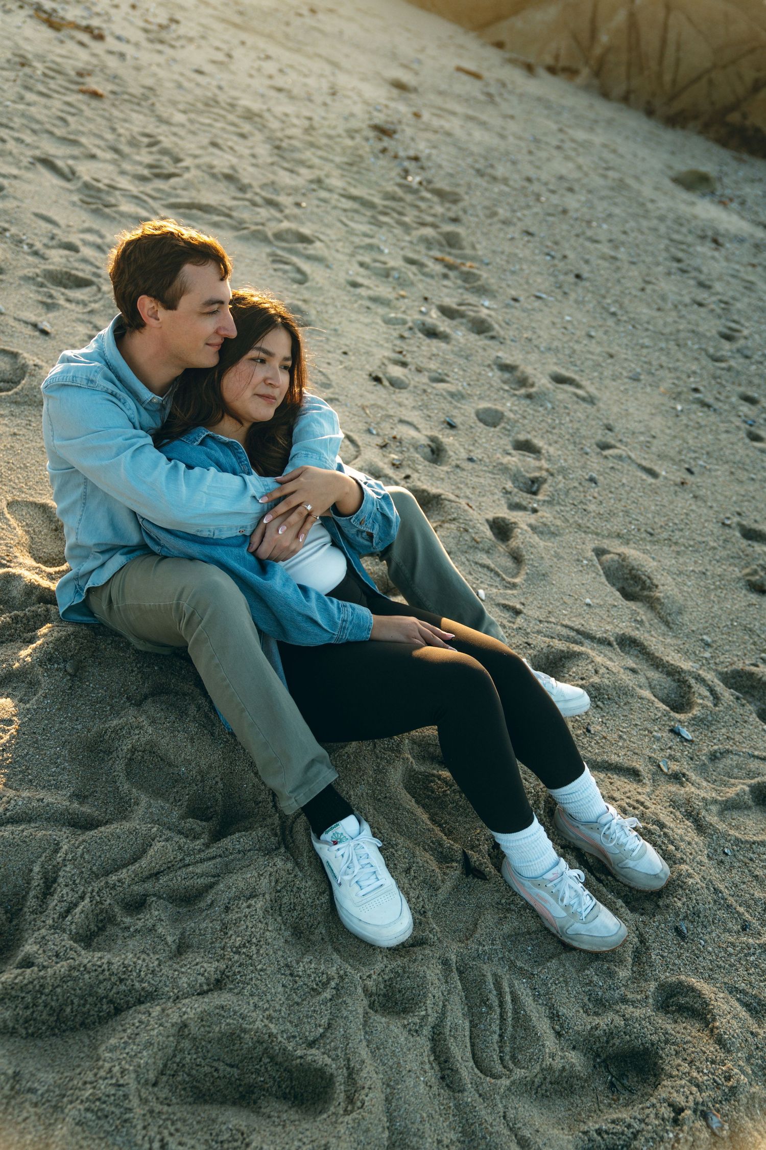 A couple sits together on a sandy beach at sunset wearing casual clothing and white sneakers.