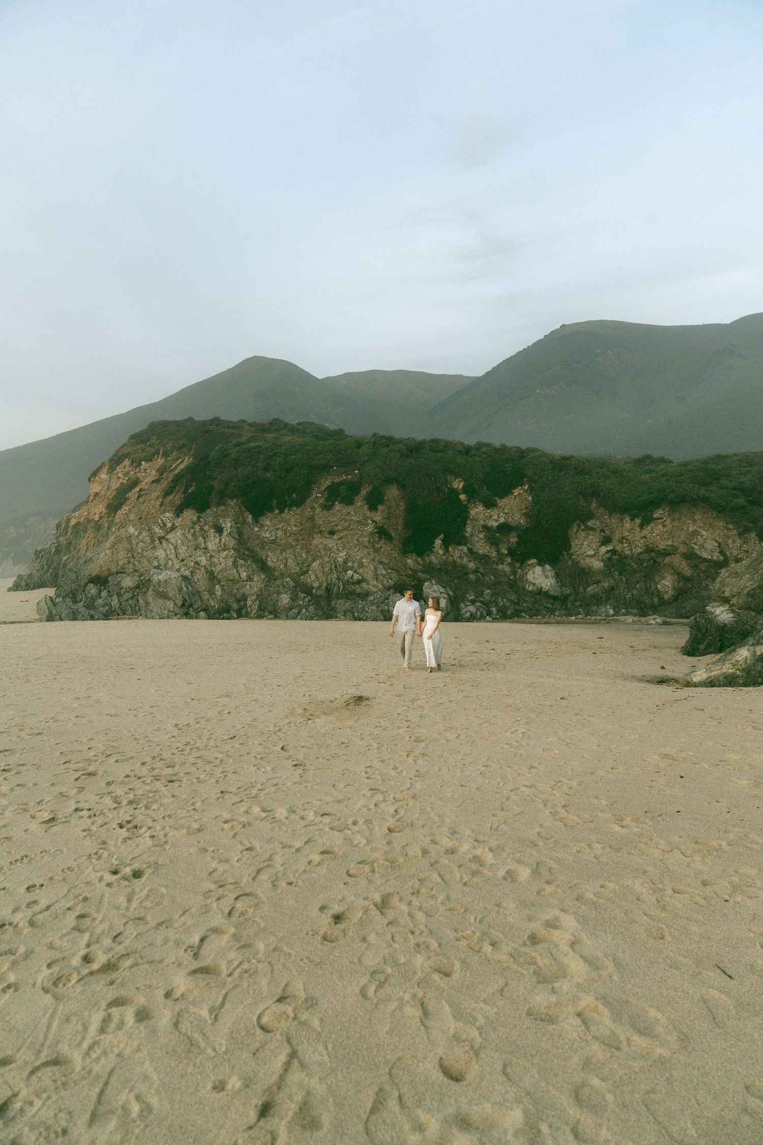 Two figures in white stand on a sandy beach with dramatic cliffs and green mountains in the background.