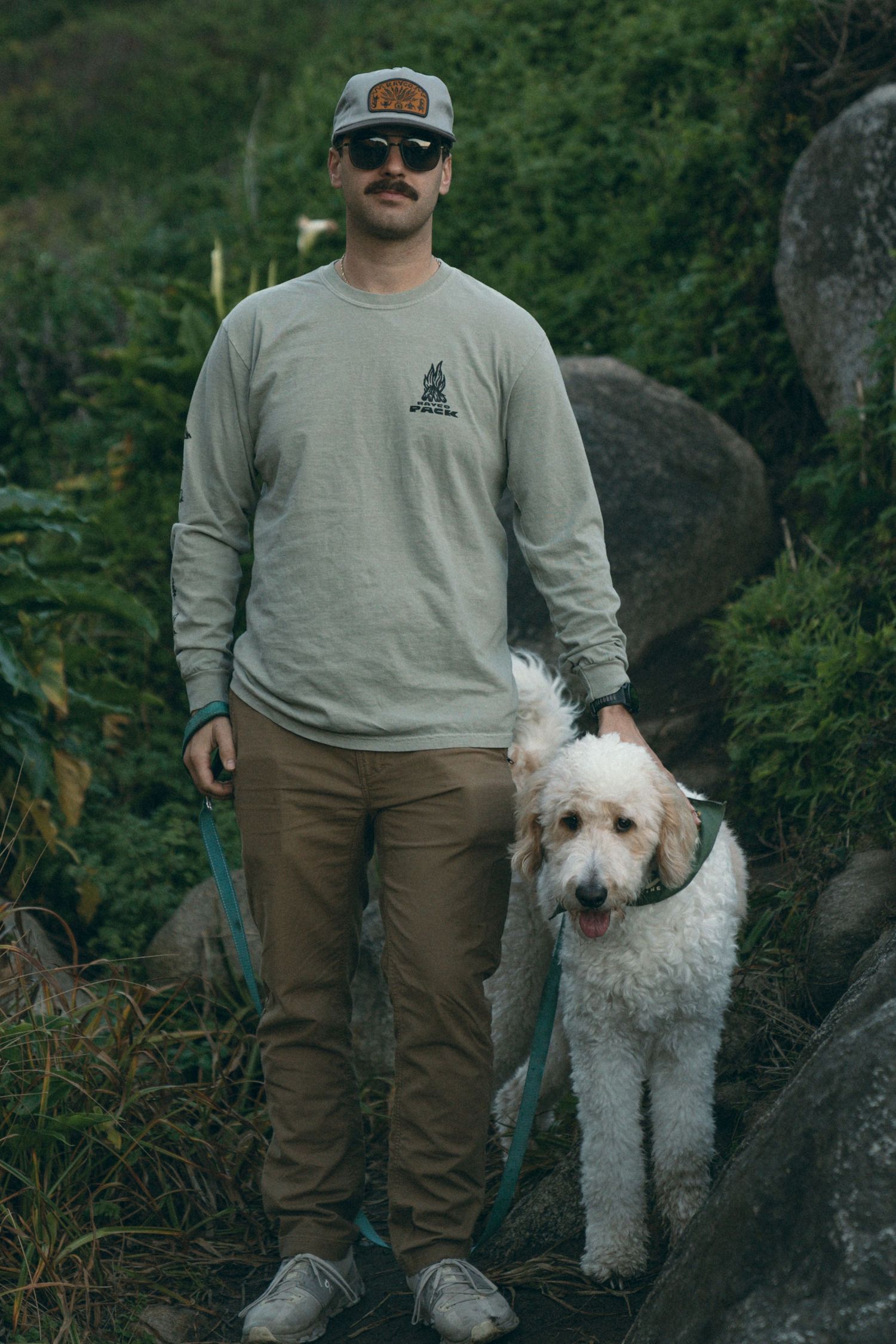 Person in gray sweatshirt and cap poses with their white fluffy dog among rocky terrain.