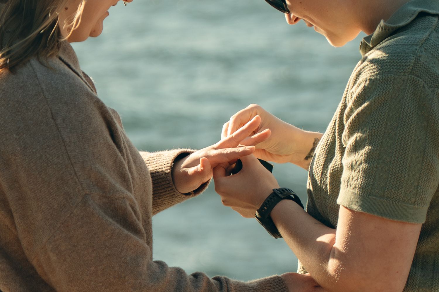 Two people holding smartphones while sitting outdoors near a body of water on a sunny day.