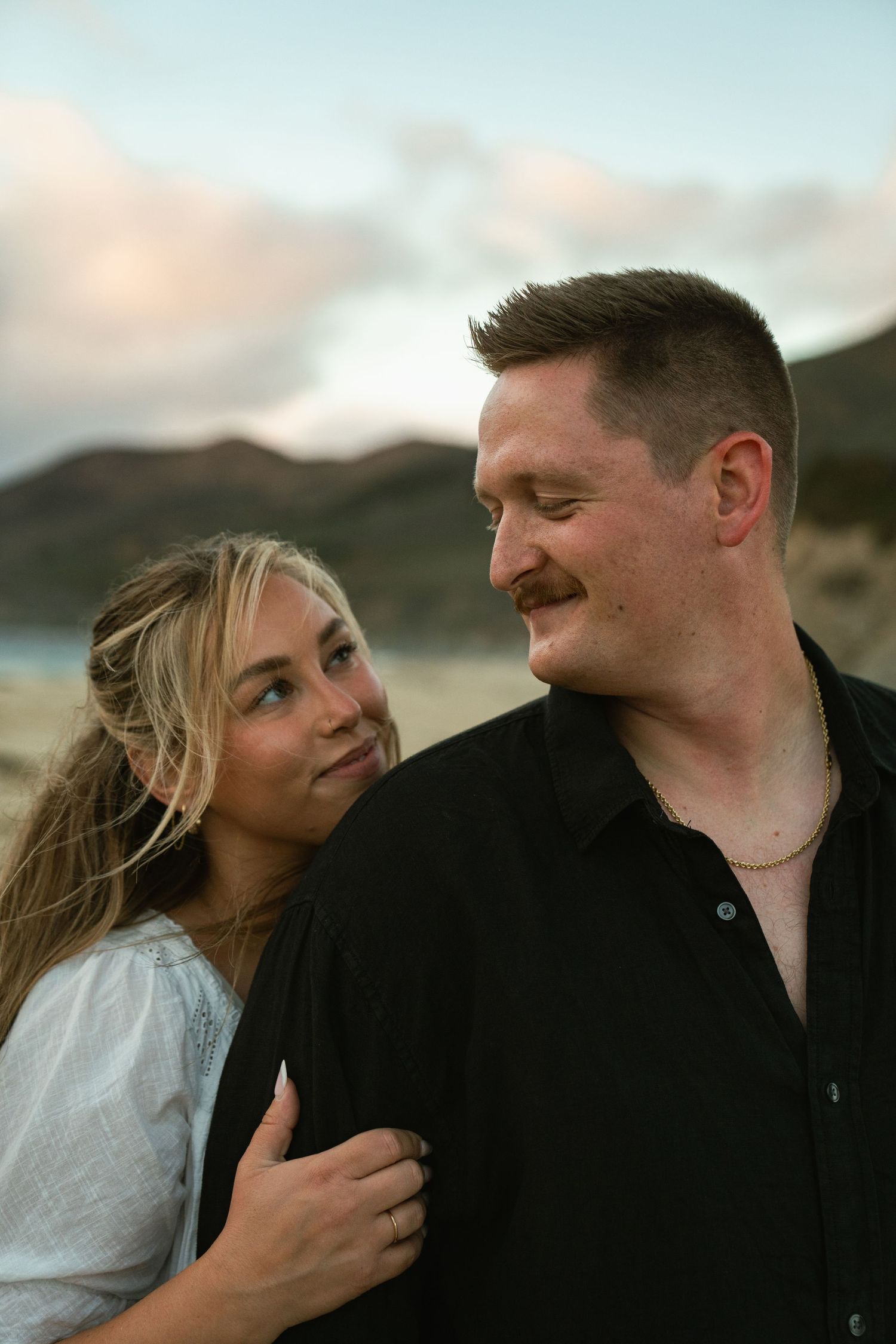 A couple shares an intimate moment at sunset with mountains and clouds in the background.