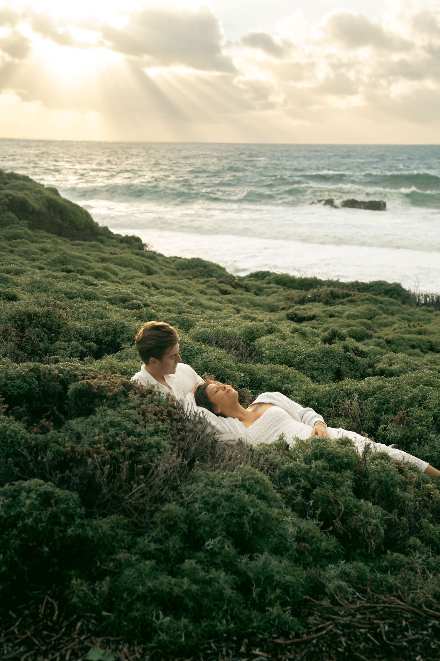 Couple in white clothing lying together in coastal greenery with ocean waves and golden sunset rays in the background.