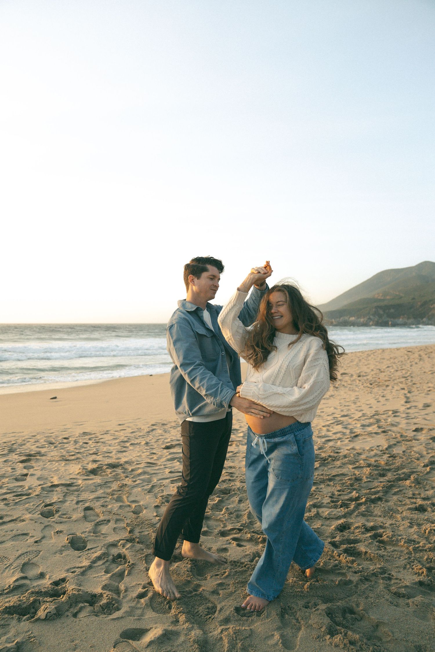 Happy couple dancing barefoot on sandy beach at sunset with ocean waves and mountains in background.