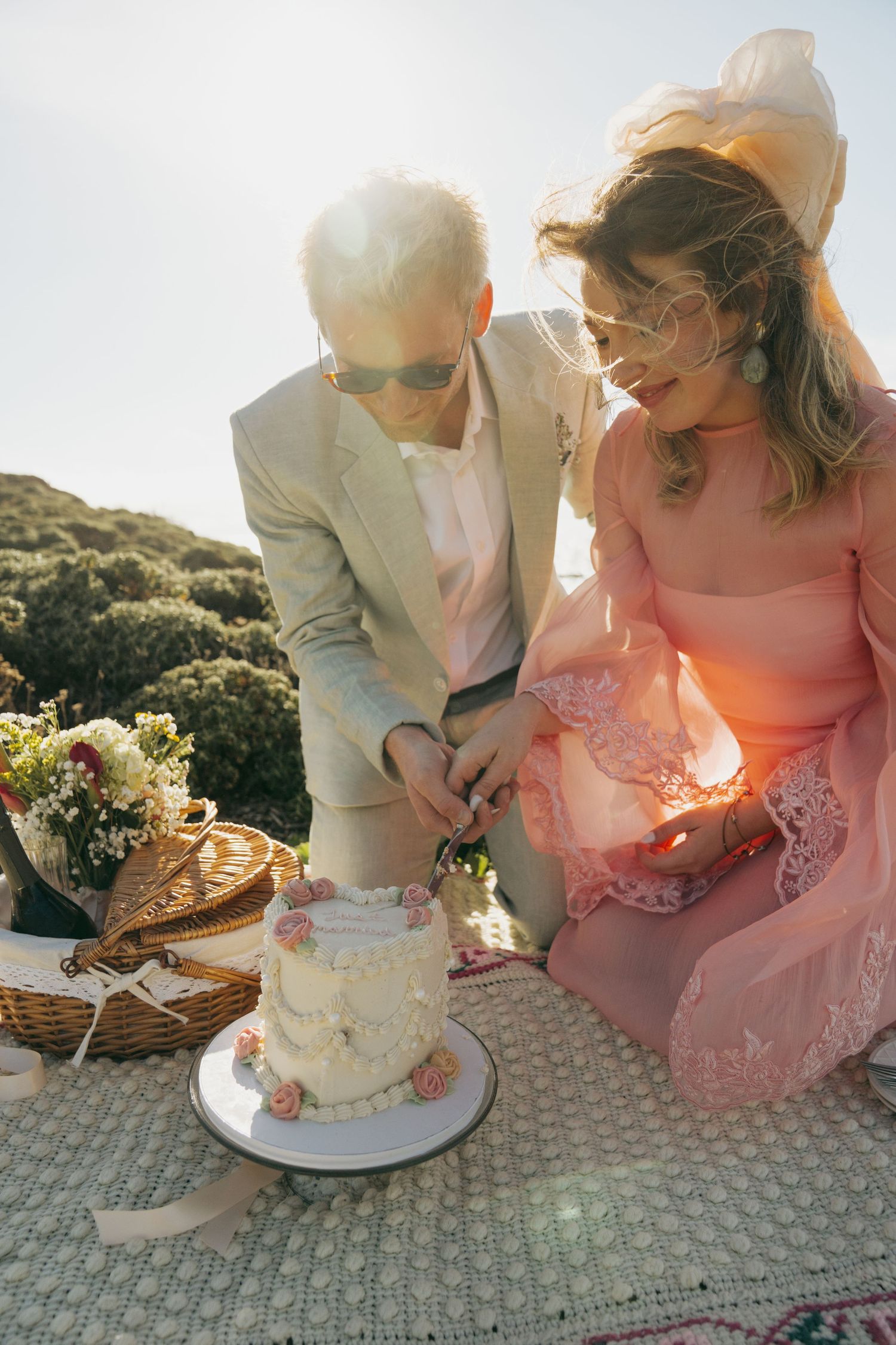 Couple in elegant attire cutting a white wedding cake outdoors at a scenic picnic setting with flowers and a wicker basket.
