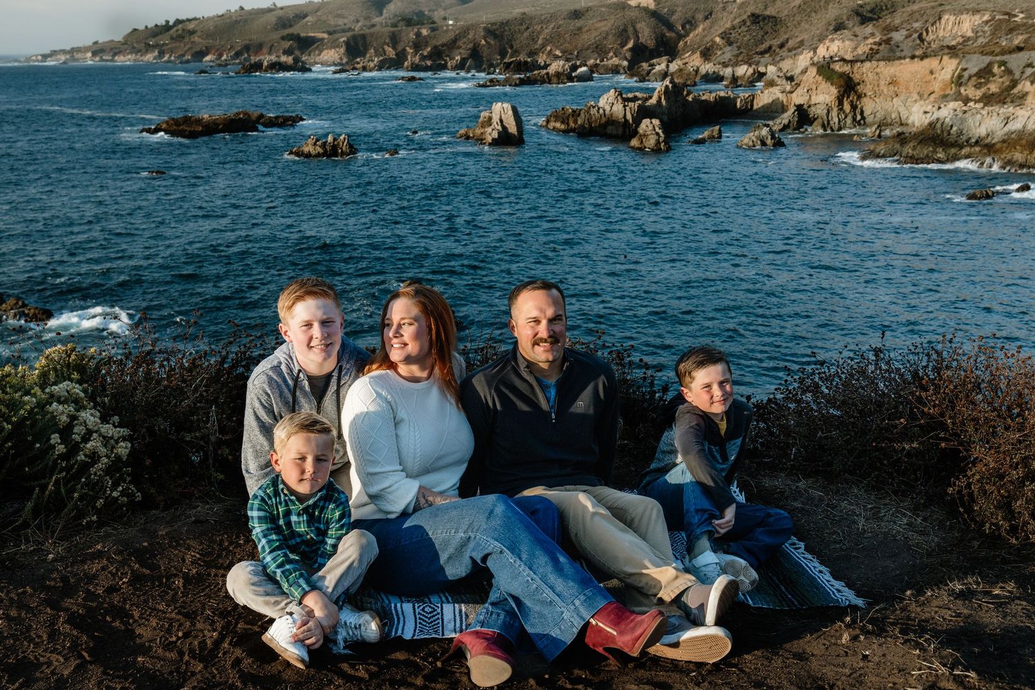 A family enjoys a scenic coastal view while sitting on a cliff overlooking the rocky Pacific shoreline on a sunny day in Big Sur.