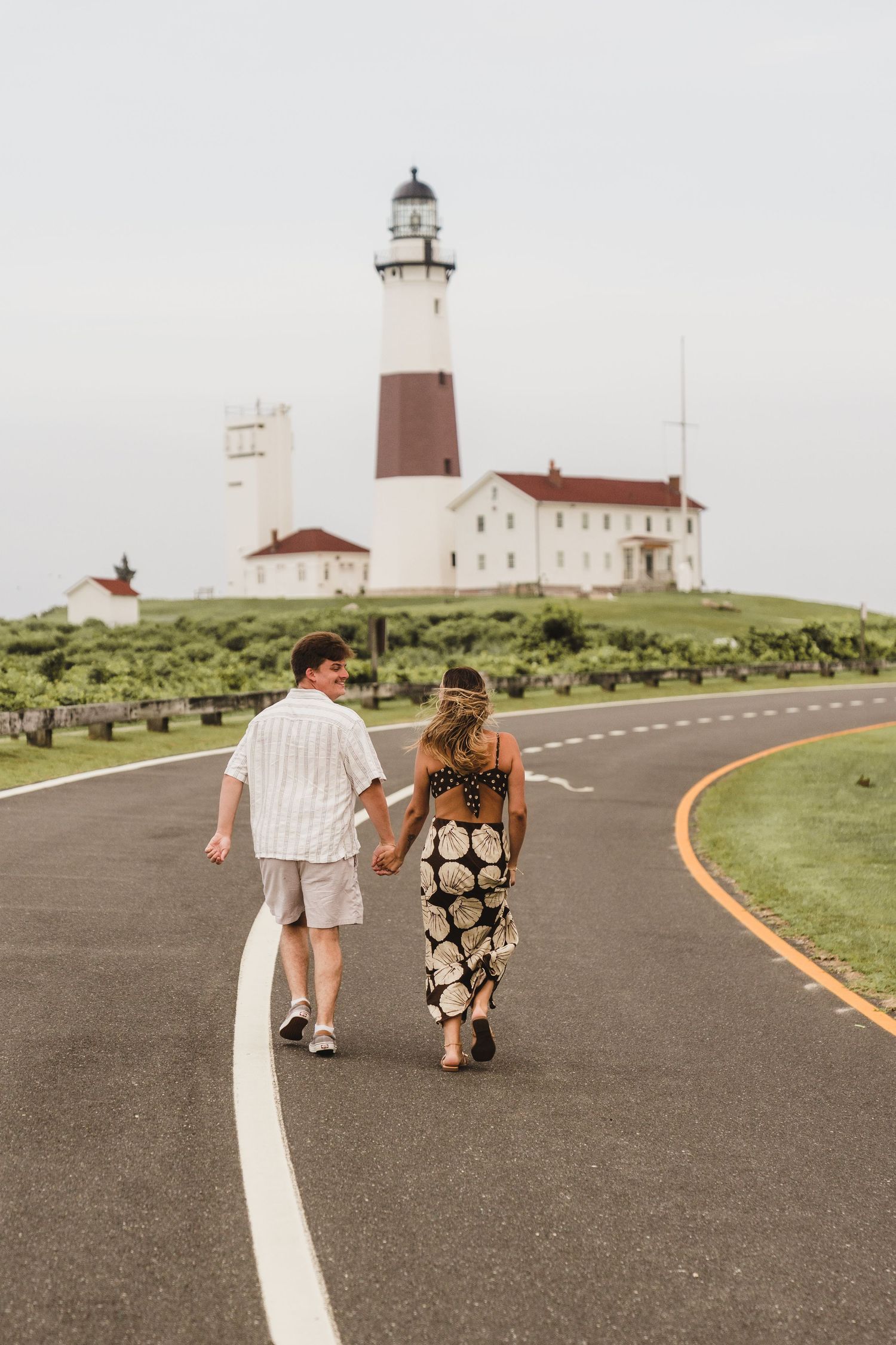 A couple walks along a curved road towards a classic white and brown lighthouse on a grassy hilltop.