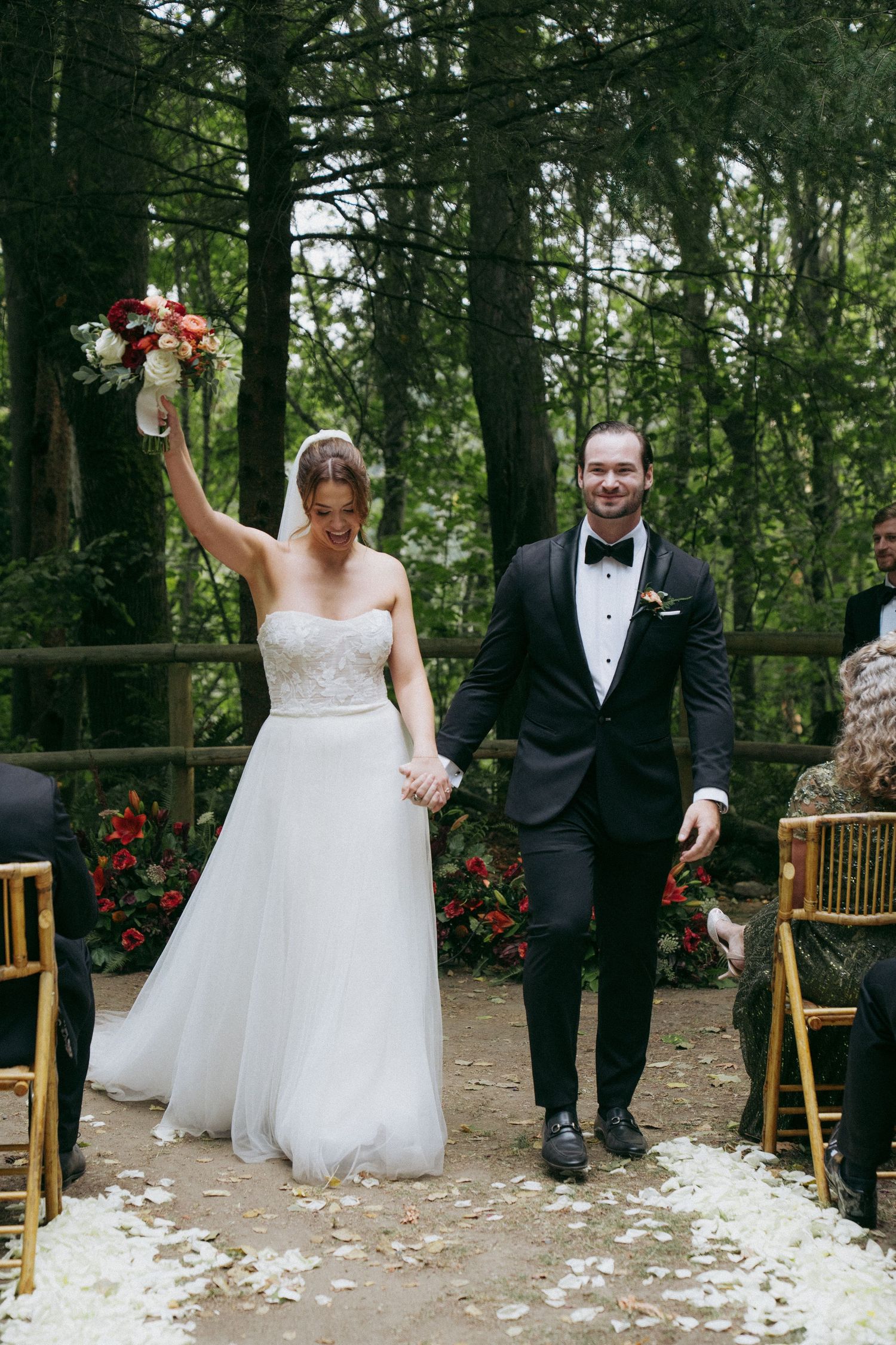 A joyful wedding ceremony exit with flower petals scattered on a forest path surrounded by evergreens.