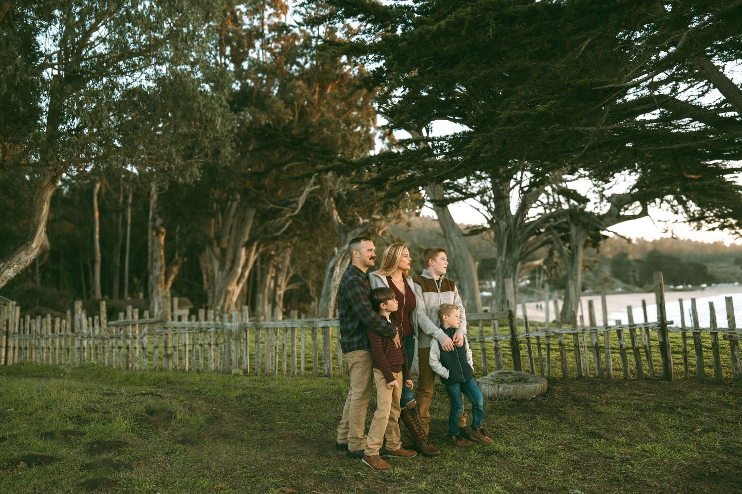 Family enjoying a stroll along a rustic wooden fence with tall cypress trees and water views in the background.