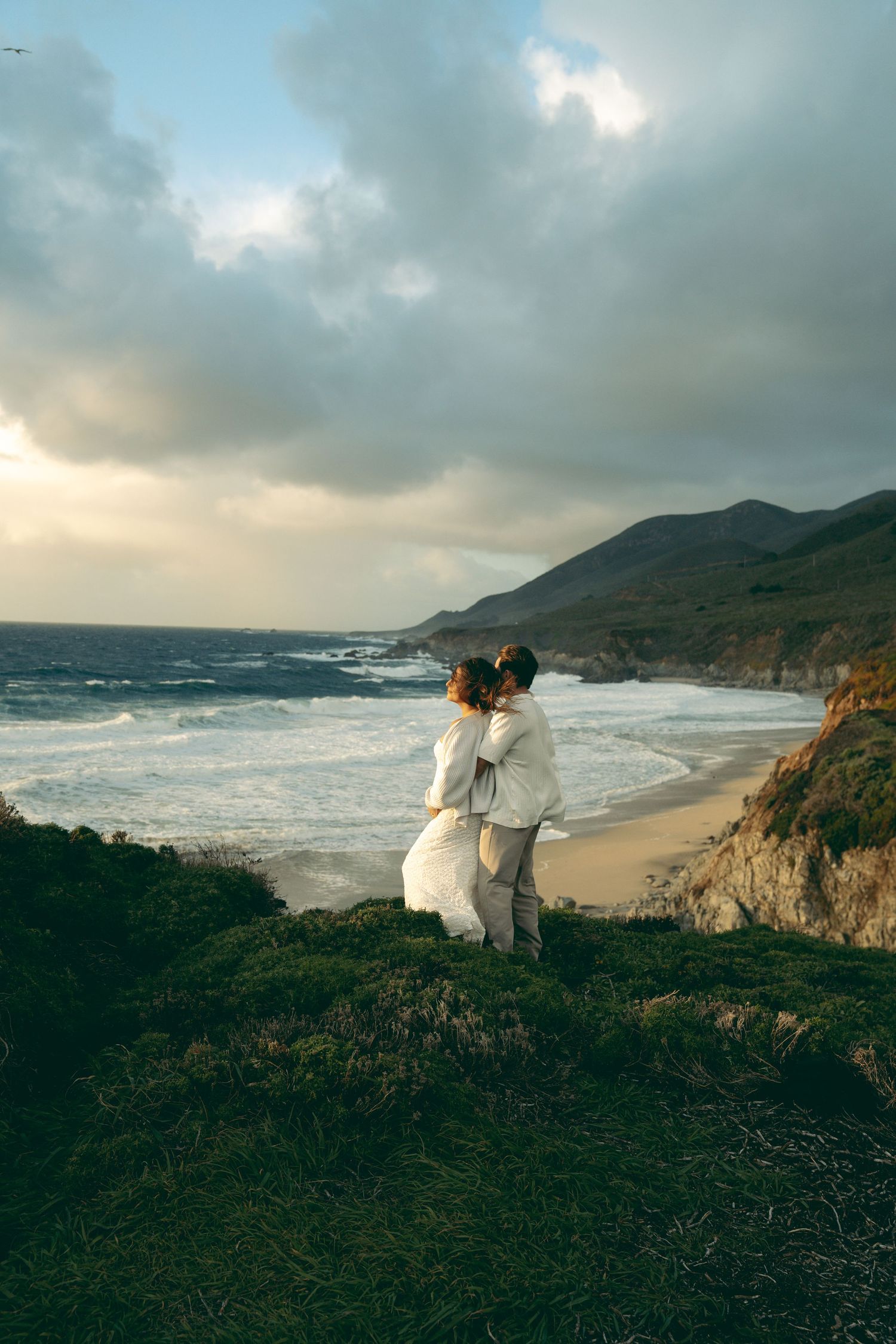 Couple embracing on a clifftop overlooking a dramatic Big Sur coastline with crashing waves and mountains at sunset.