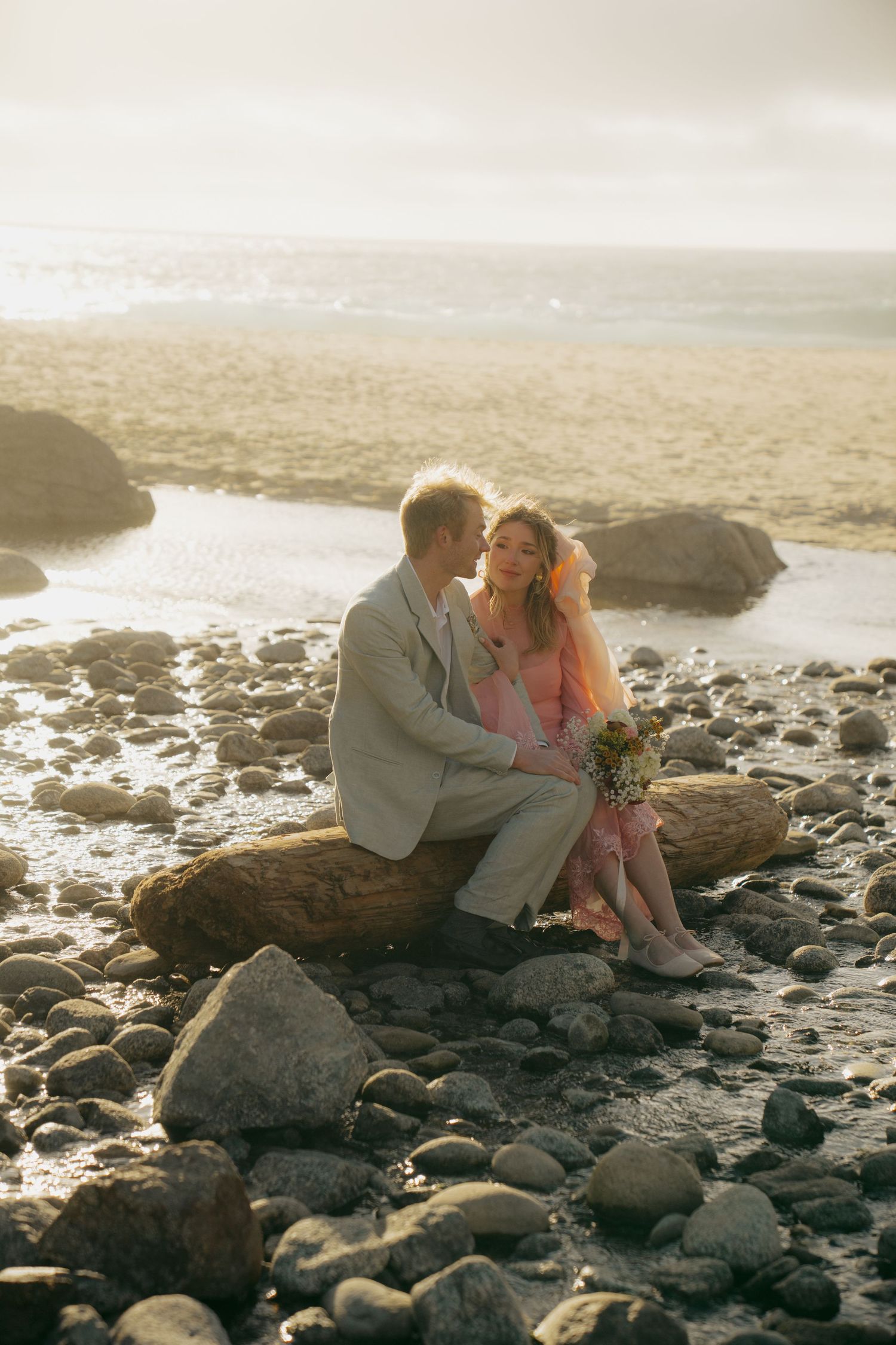 Romantic couple sitting on driftwood on a rocky beach at golden hour, woman holding a bouquet in pink dress.