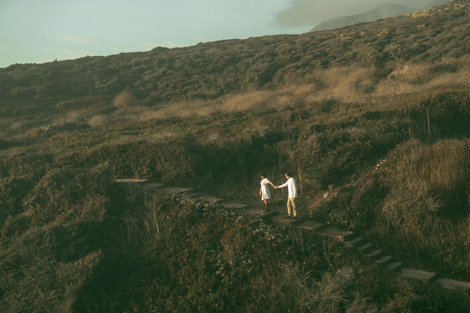Two figures in white stand on a winding mountain trail surrounded by misty brown hills and rugged terrain.