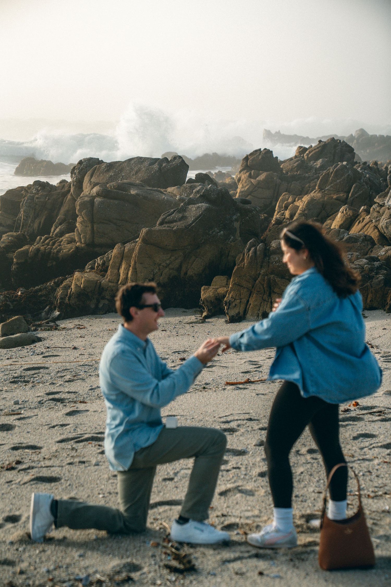 A romantic beach proposal with crashing waves and rocky coastline in the background.