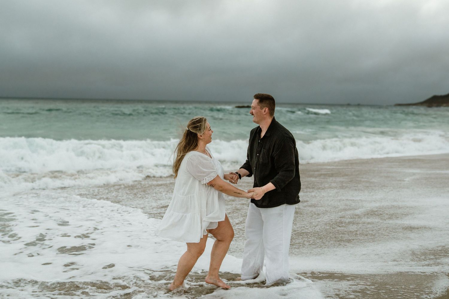 A couple dances along a misty ocean shoreline with waves crashing behind them.