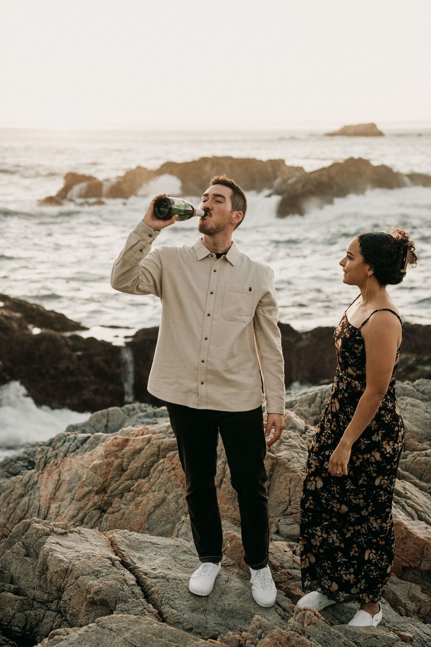 Couple enjoying a scenic moment on rocky coastal cliffs at sunset while drinking from a bottle.