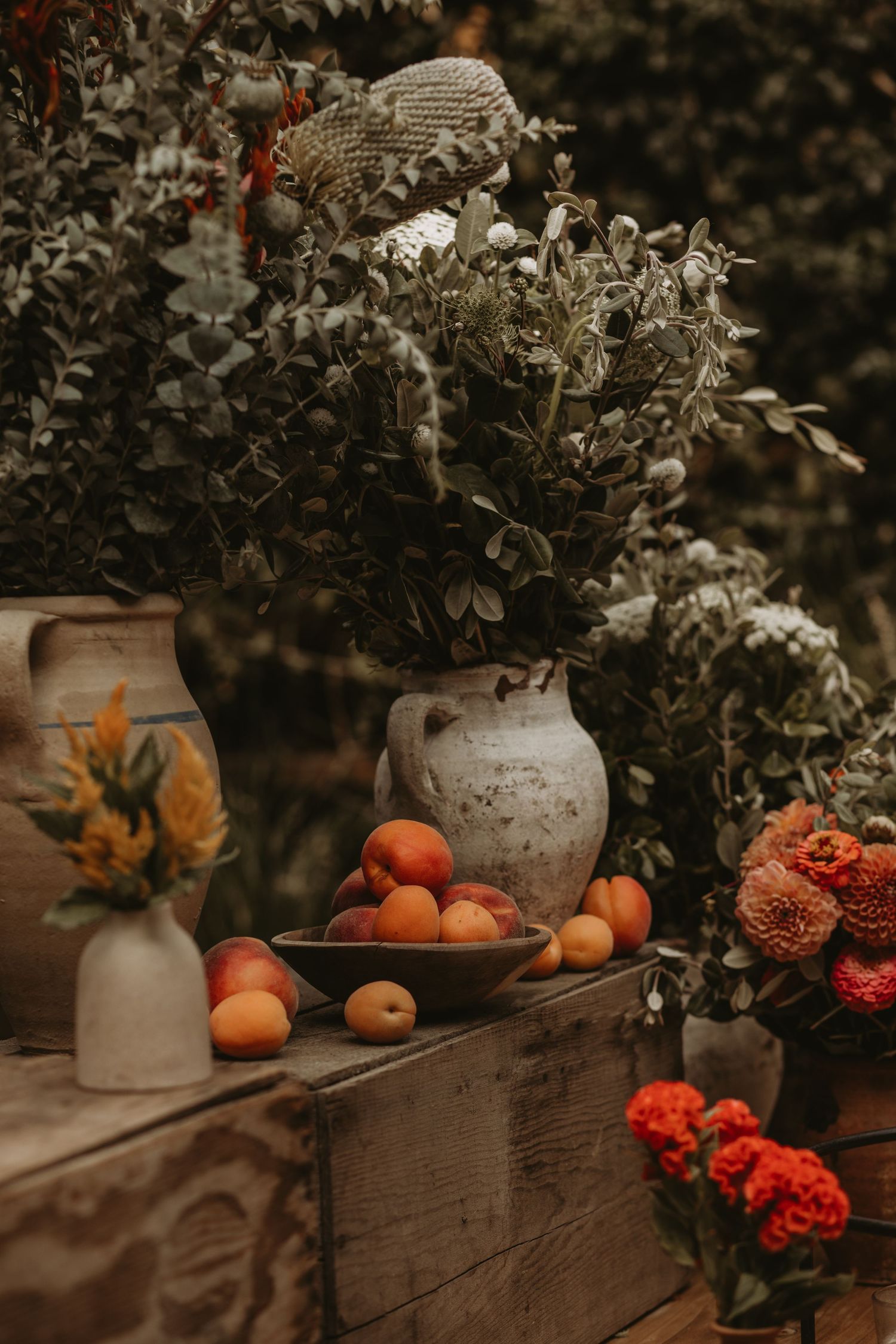 Rustic still life with fresh oranges, vintage vases, red flowers, and decorative plants in moody autumn lighting.