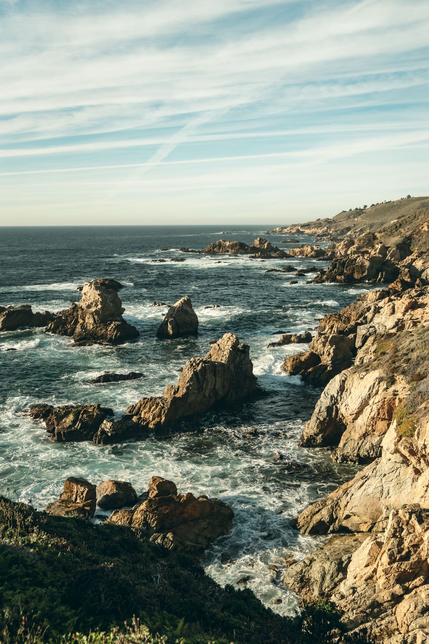 Rugged rocky coastline with crashing waves against dramatic cliffs under a bright blue sky.