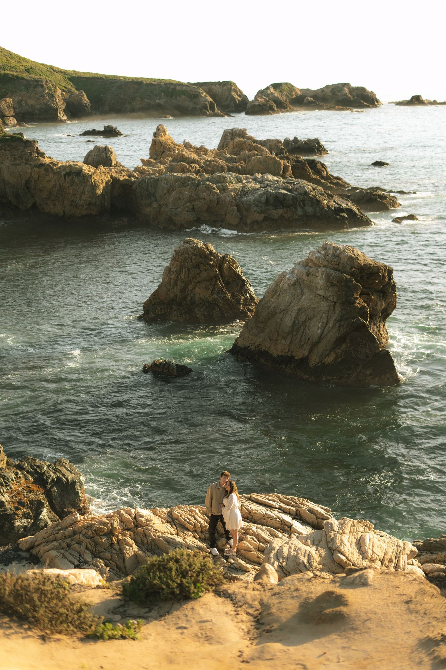 Couple embracing on rocky coastal cliffs with turquoise ocean waters and jagged sea rocks in the background.