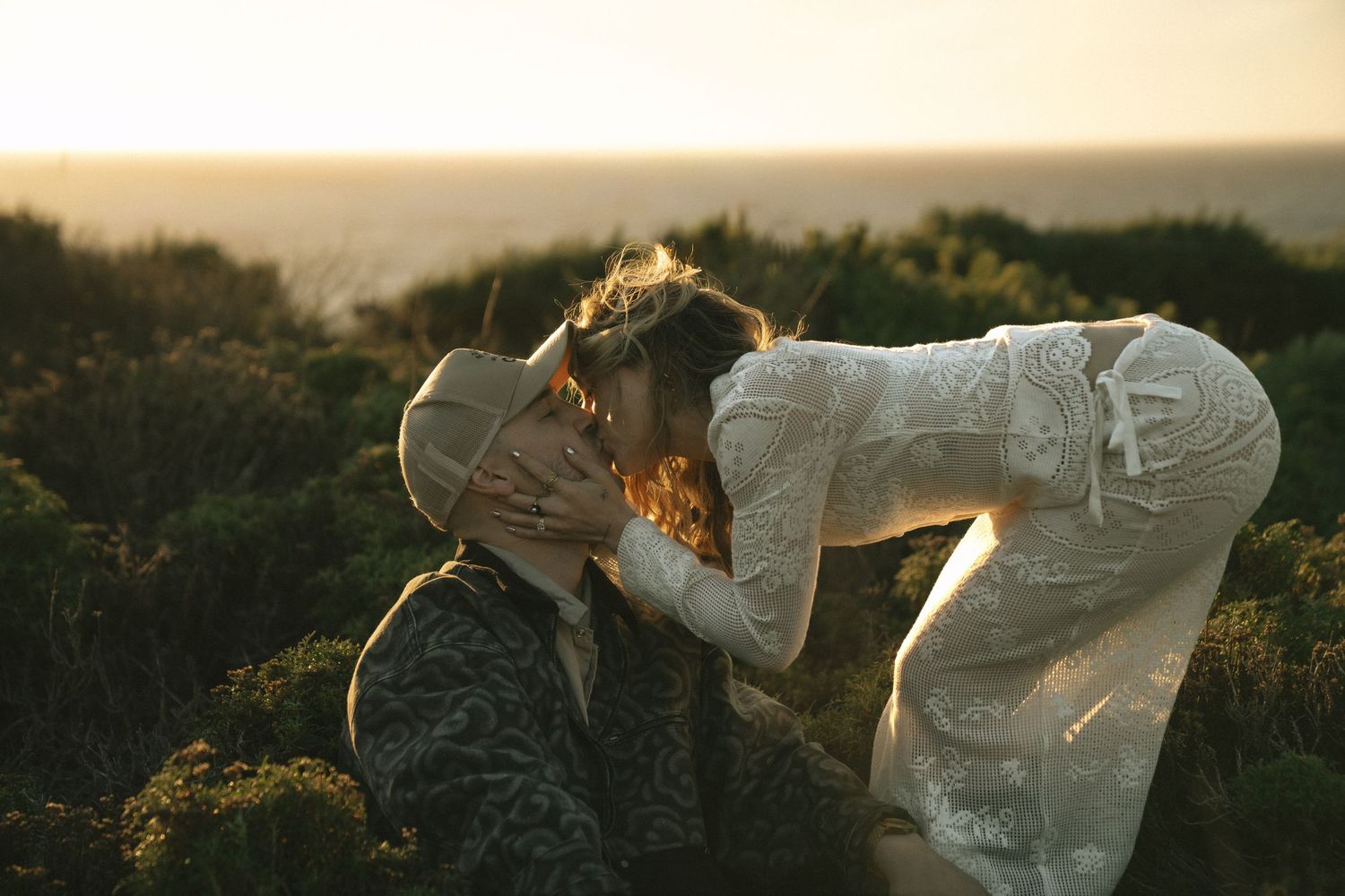 Couple sharing a romantic kiss at golden hour with ocean views and lush greenery in the background.
