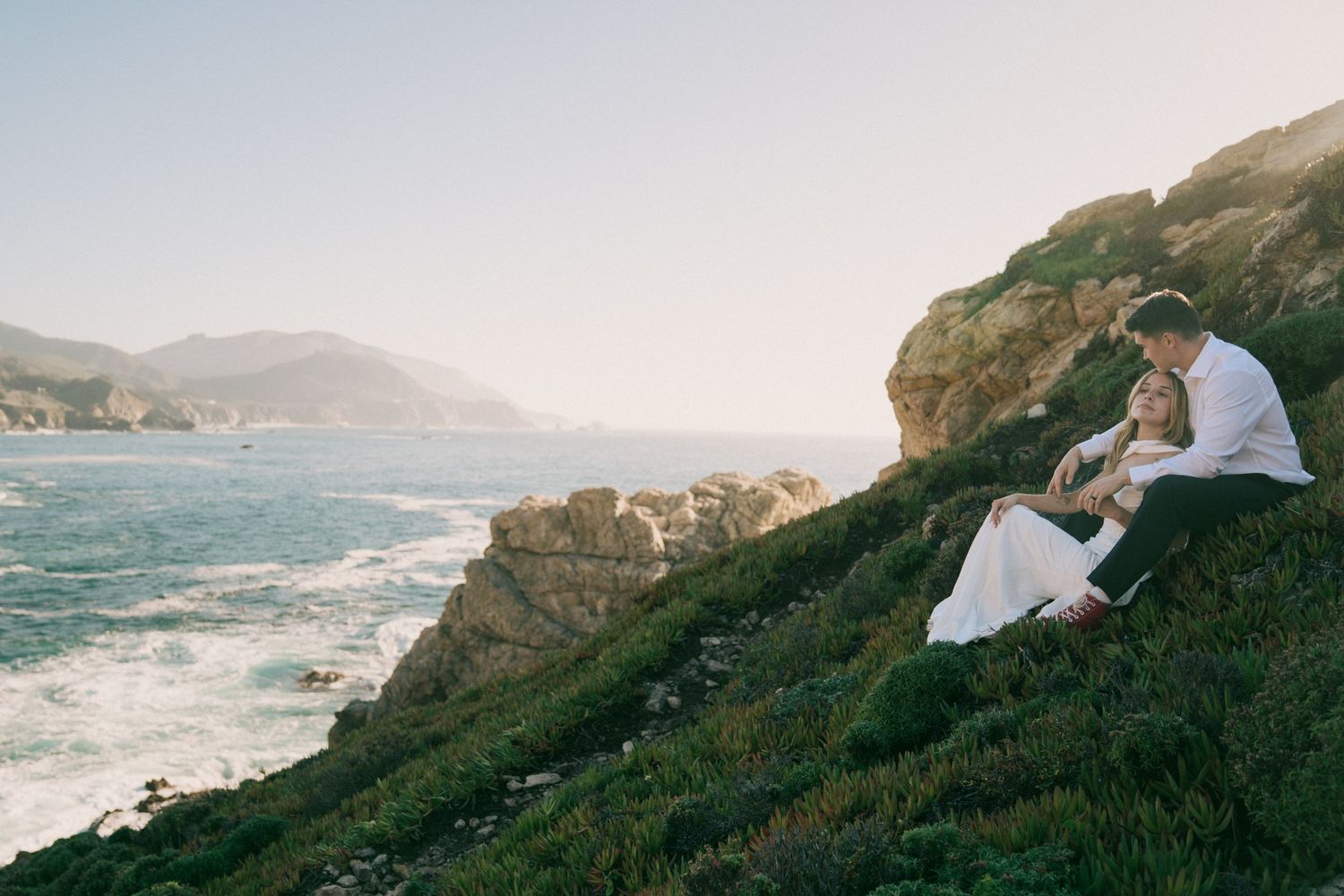 A couple sits on a coastal cliff overlooking the rugged Pacific Ocean shoreline at sunset.
