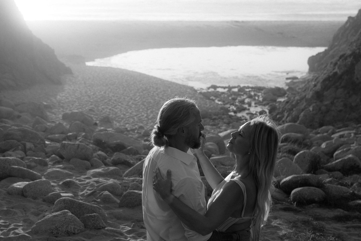 Two people embrace while sitting on rocky coastal cliffs overlooking a misty ocean landscape at sunset.