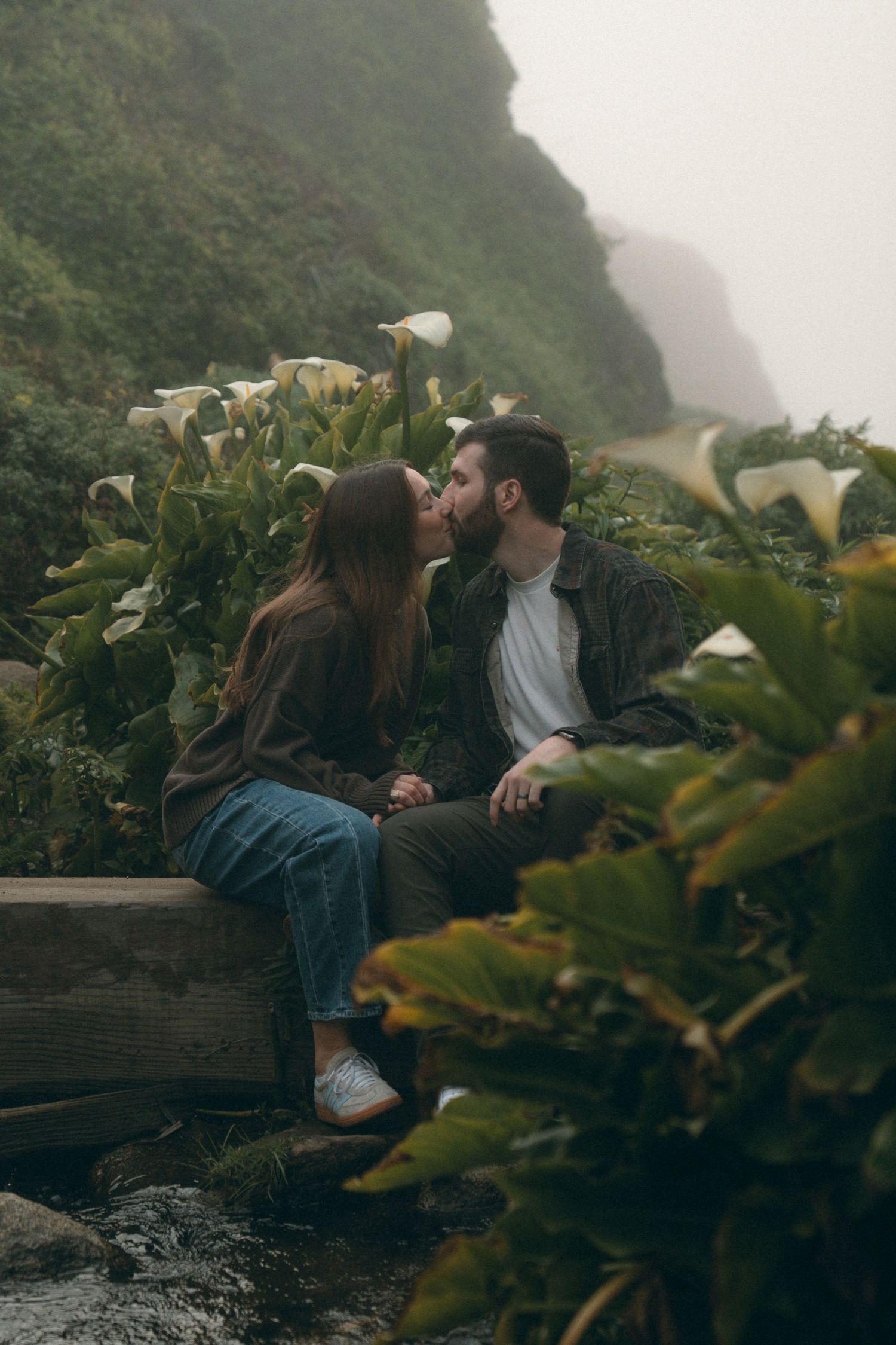 A couple sharing an intimate moment on a wooden bench surrounded by calla lilies and mountain views.
