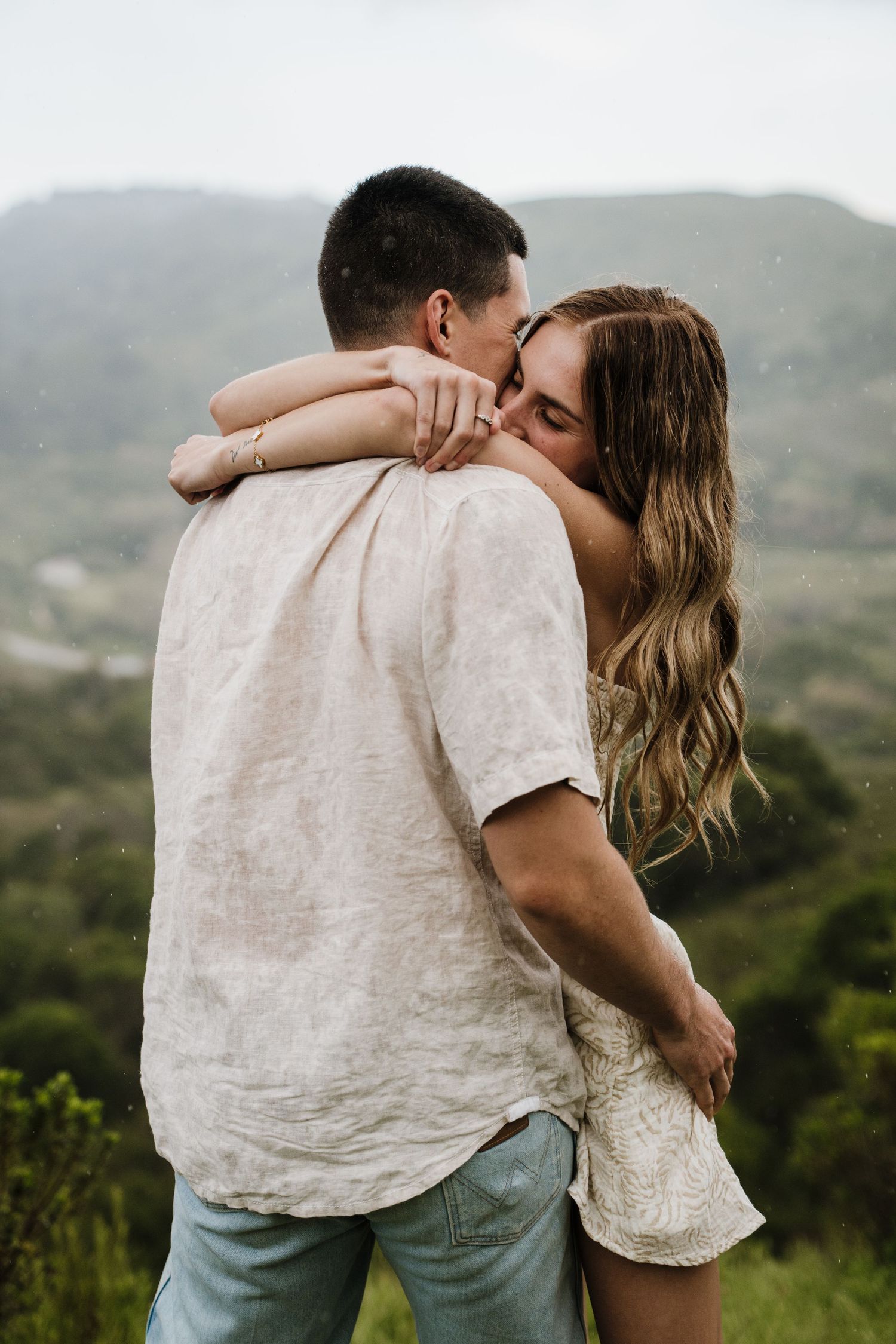 A couple shares a tender embrace against a backdrop of lush mountain scenery.