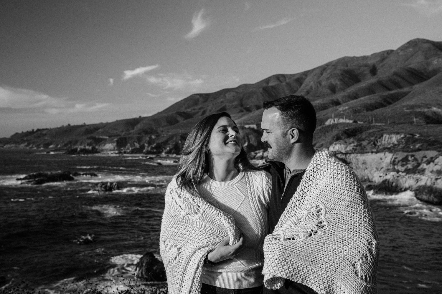 A romantic black and white portrait of a couple in sweaters embracing along a rocky coastal landscape with mountains.