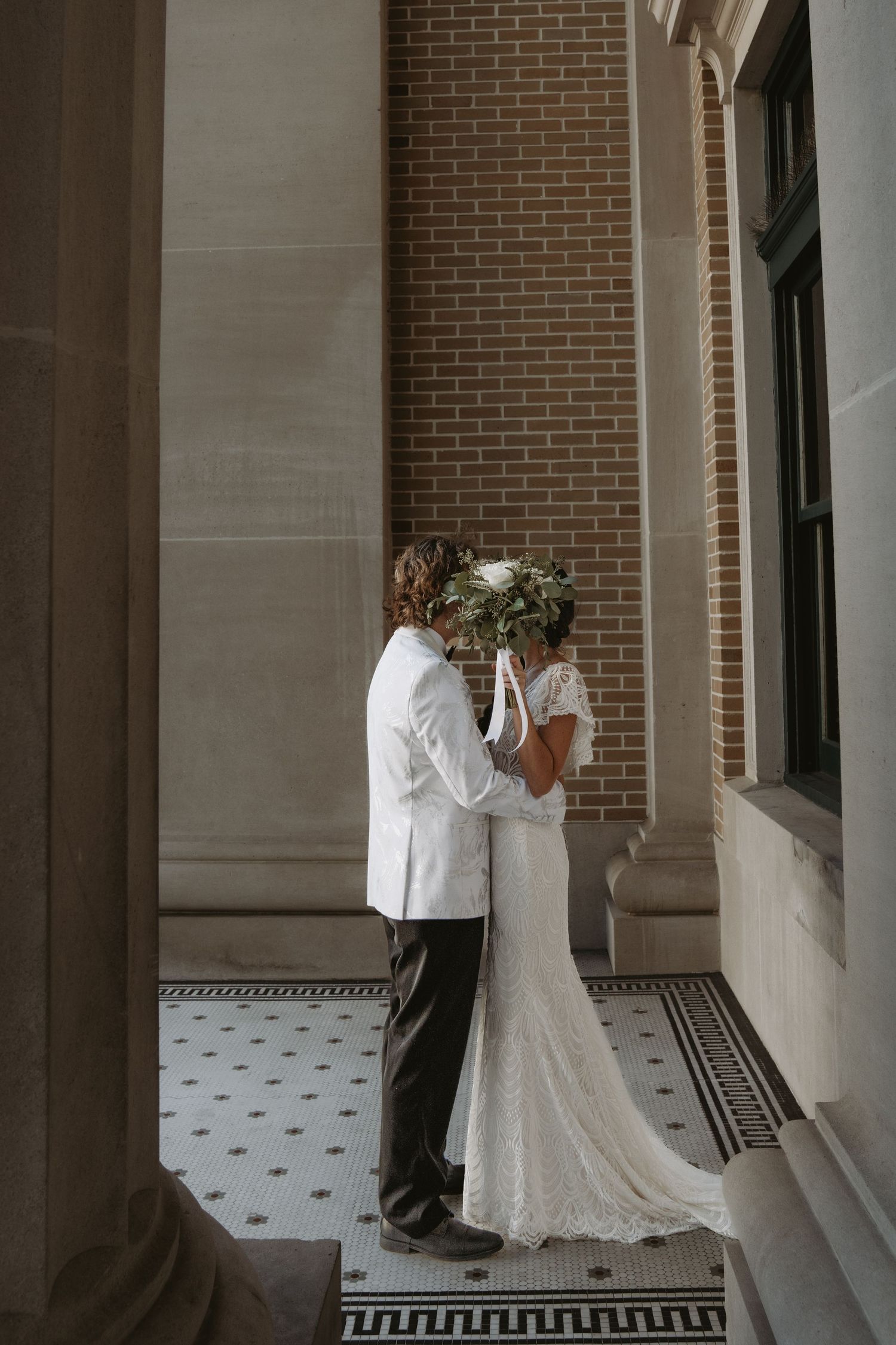 A couple in wedding attire embraces on an elegant portico with brick walls and tiled floor with Greek key pattern.