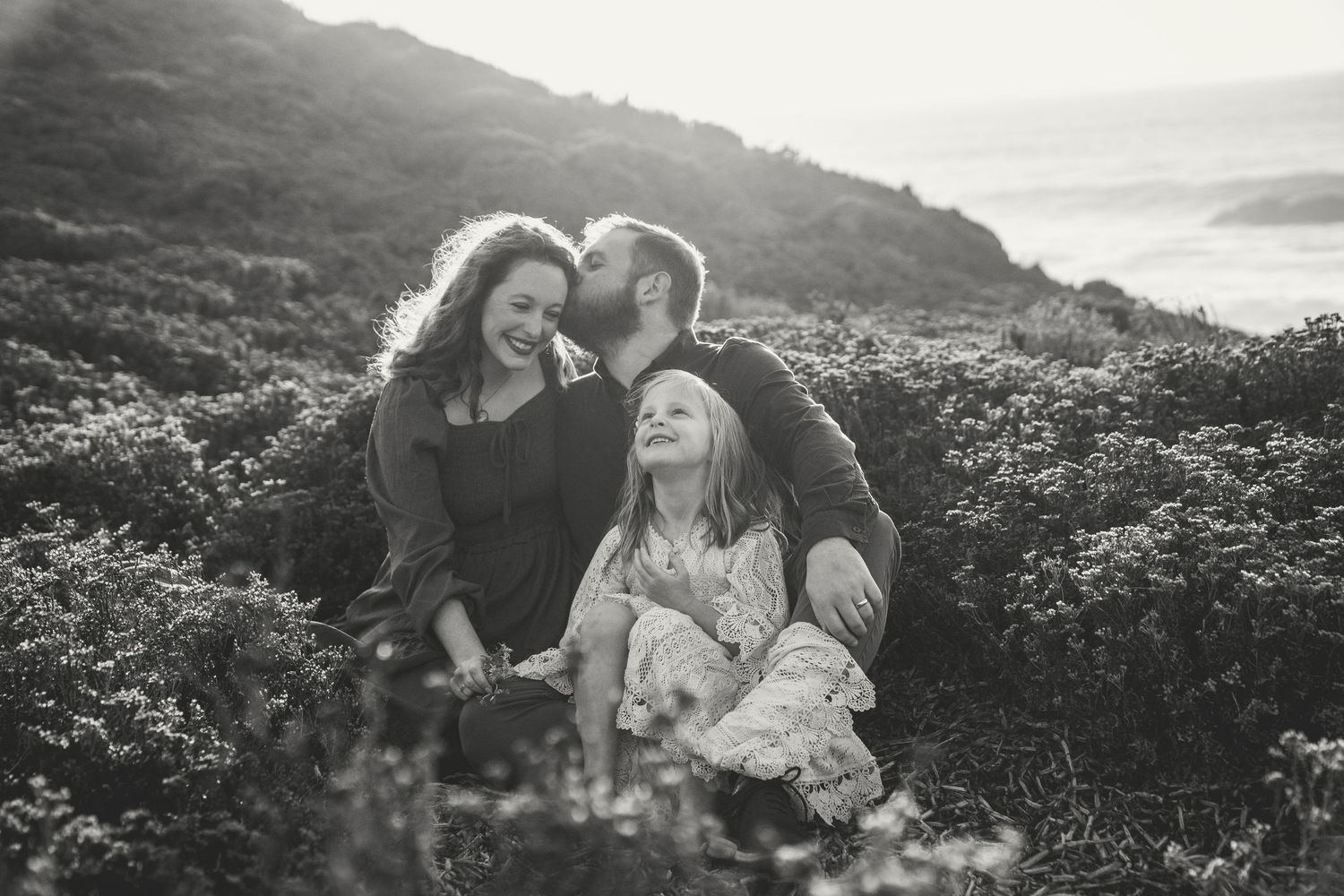 A loving family sits together among wildflowers on a hillside overlooking a misty valley in black and white.