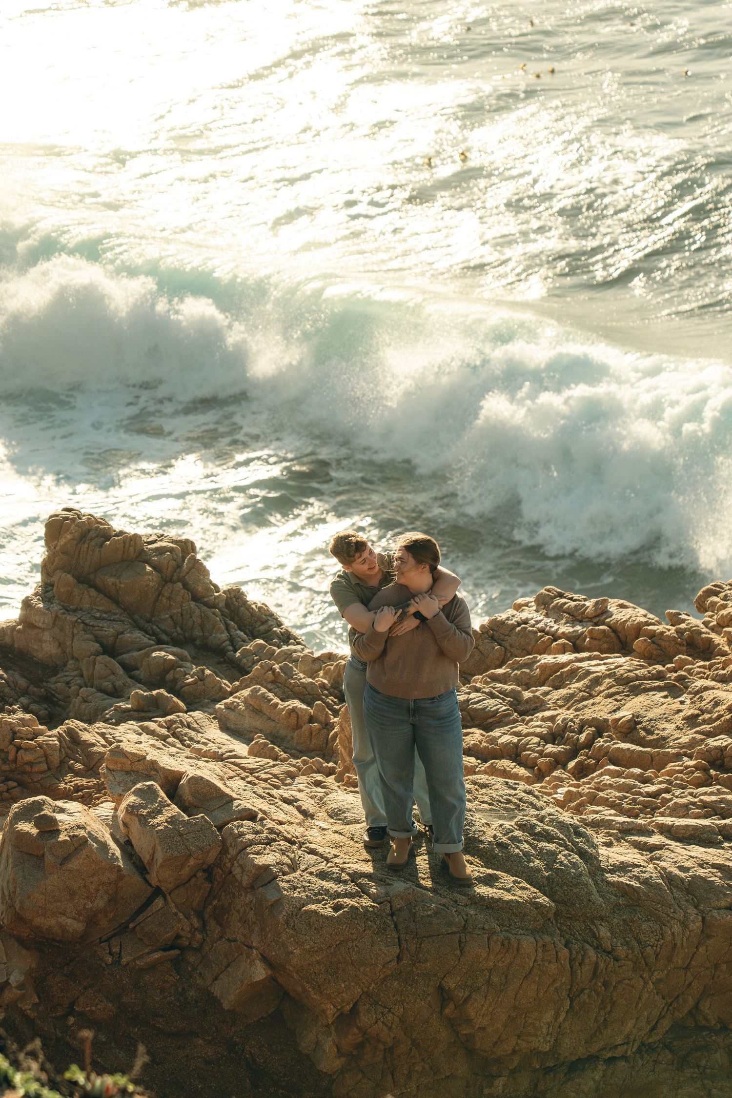 Couple embraces on rocky coastal cliffs with crashing ocean waves in the background at sunset.