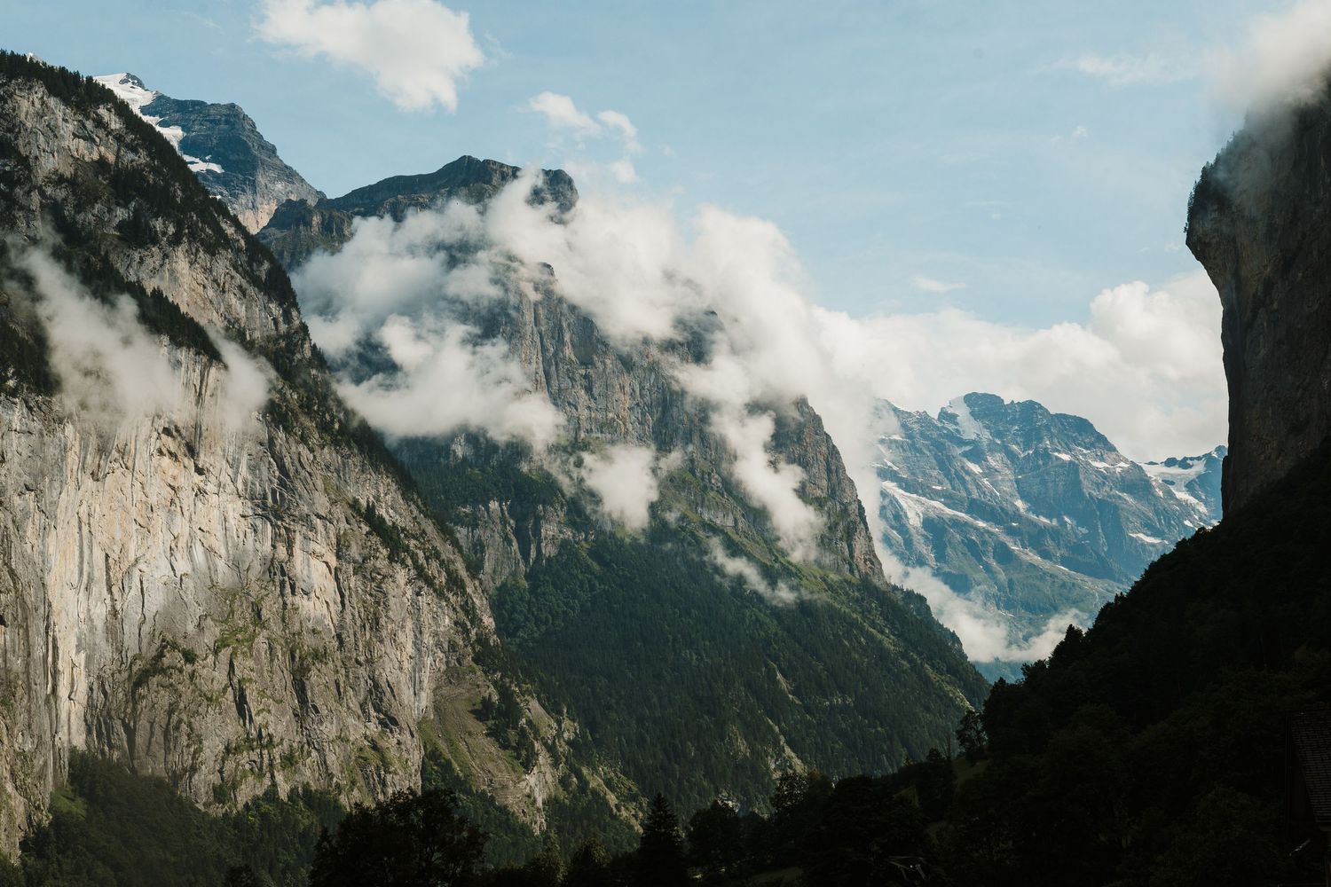 Dramatic mountain peaks with low-hanging clouds in a valley surrounded by steep rocky cliffs on a sunny day.